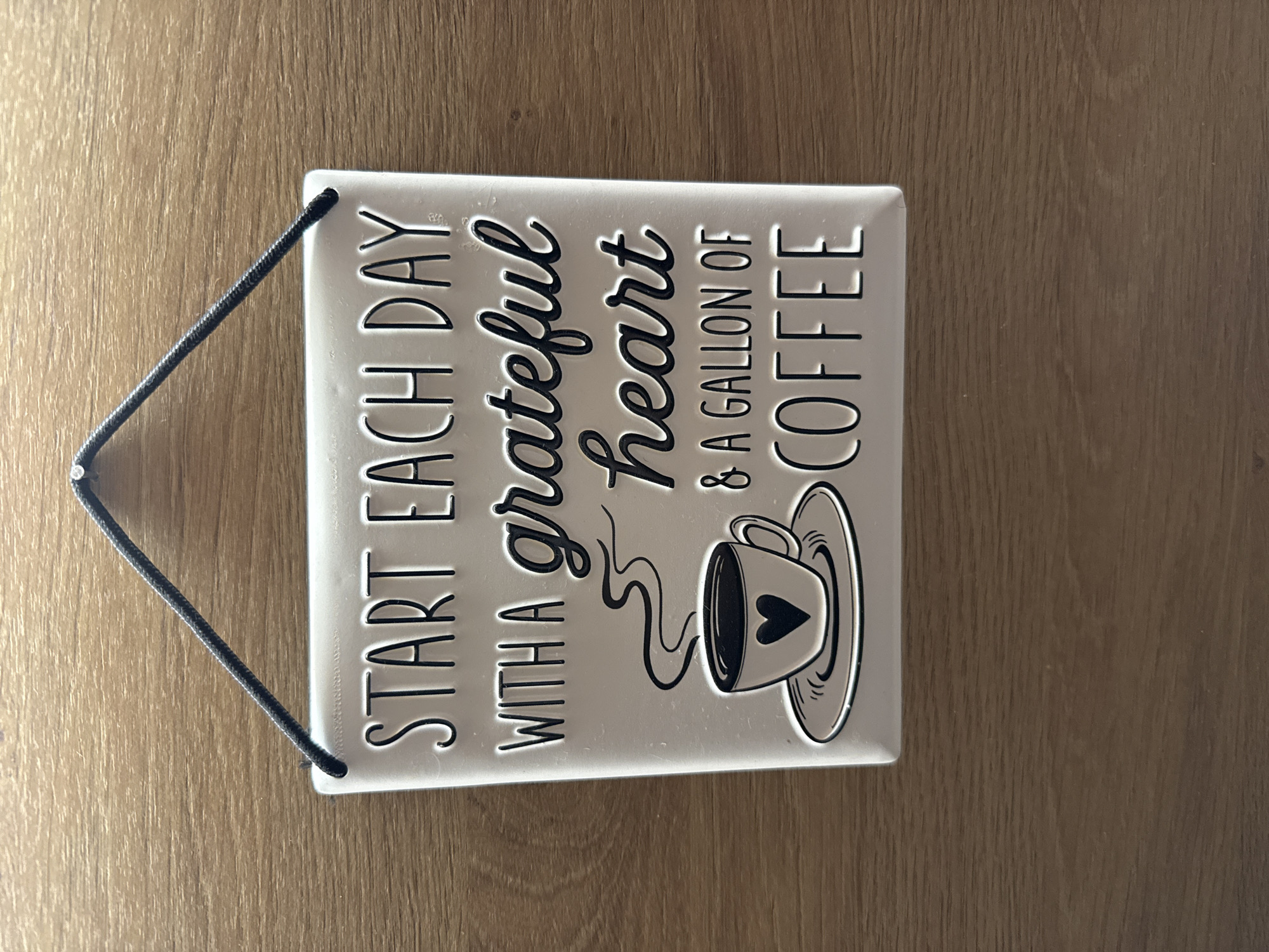 A rectangular, light-colored sign or plaque is horizontally displayed on a wooden surface. The sign features black text and a graphic against a white or light grey background. A black braided cord is attached to the upper left and upper right corners, forming a triangular hanger.
The text on the sign reads: "START EACH DAY WITH A grateful heart & A GALLON OF COFFEE." The words "grateful" and "heart" are rendered in an elegant, flowing script font, while the remaining text is in a clear, bold, sans-serif typeface. Below "heart," there is a black outline illustration of a steaming coffee cup, containing a small heart shape, resting on a saucer. The text and illustration appear to be slightly raised, giving them a three-dimensional quality.
The sign rests on a warm, medium-brown wooden surface with a distinct vertical grain pattern, suggesting a table or floor. Bright, possibly overhead, lighting creates reflections and a subtle glare on the sign's glossy surface. No individuals are present, and no activity is depicted beyond the presentation of the sign itself. The message conveys a positive and appreciative sentiment, linking gratitude with a love for coffee. The setting is an interior space, consistent with a home or cafe in Sofia, Bulgaria.