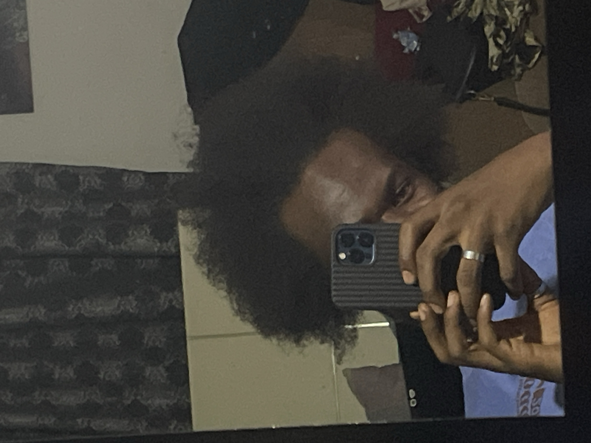 The image depicts a person taking a selfie in a mirror. The individual has dark skin, and dark, curly hair. They are holding a smartphone with a grey case, capturing their reflection. The person is wearing a blue shirt. The reflection also shows a glimpse of what seems to be clothing in the background. The mirror is surrounded by what appears to be a wall with patterned wallpaper. The location of the image is not explicitly identified as Kuchingoro, Nigeria.