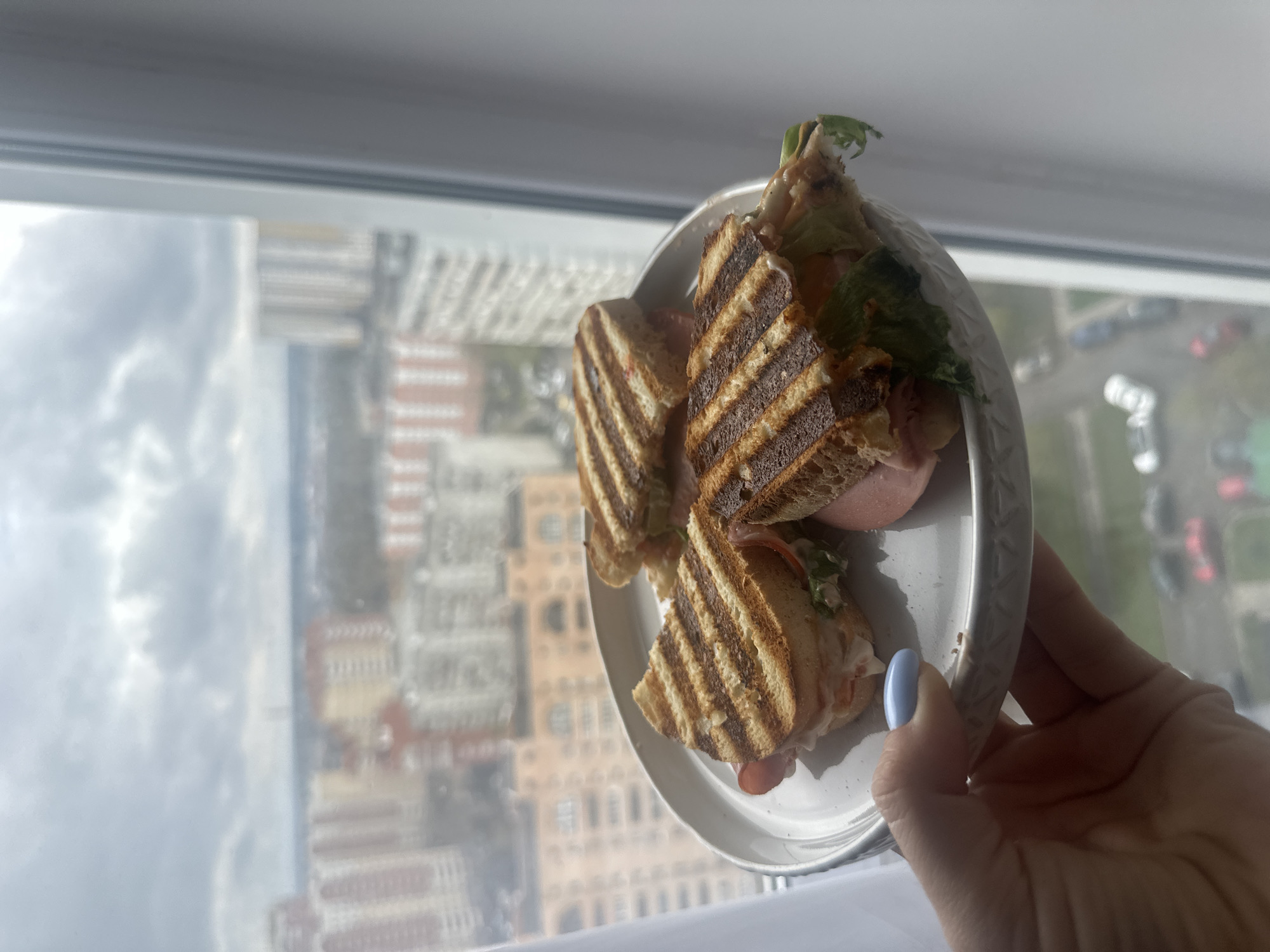 A person holds a white plate with three halved, grilled sandwiches against a backdrop of Gavrilkovo, Russia's urban landscape. The sandwiches appear to contain fillings of lettuce, tomato, ham and sauce, with a bread that is striped by grill marks. The buildings of Gavrilkovo are visible through a window, with the sky overcast. The person's hand, painted with a light blue nail polish, is clearly holding the plate.