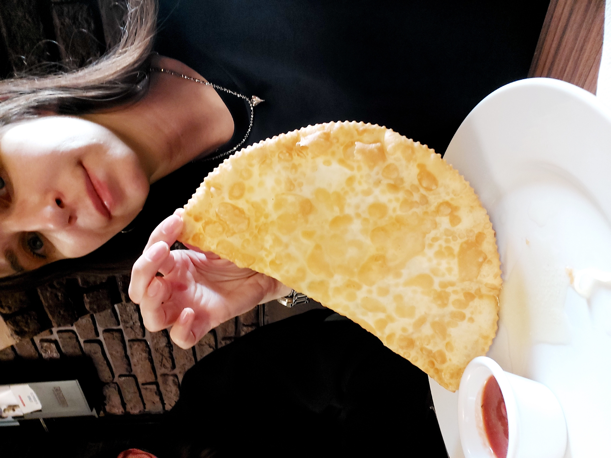 A person holds a large, deep-fried pastry resembling a cheburek, near a small white cup containing red sauce, on a white plate. The pastry has a golden-brown, bubbly surface. The person has dark hair and is wearing a dark top. The scene appears to be indoors, possibly in a restaurant. Based on the style of food, this image is likely located in the city of Saint Petersburg, Russia.