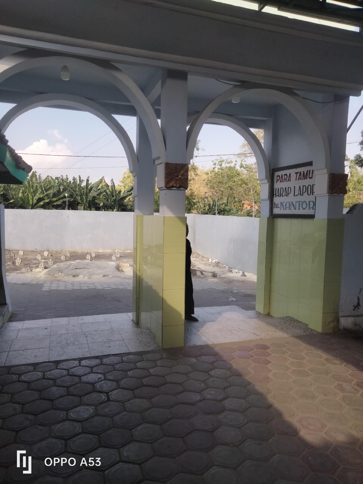 The image depicts an outdoor scene in what appears to be a public facility or administrative building in Badung, Indonesia. The foreground is dominated by a pathway paved with hexagonal interlocking bricks, casting distinct shadows. To the left, a lighter-colored tiled area is visible, leading towards an entranceway.
The entranceway is characterized by a series of light blue arched structures supporting a ceiling. On the right side of the entrance, a sign is mounted on a pillar, displaying text in Indonesian. The visible portion of the sign reads "PARA TAMU HARAP LAPOR ke KANTOR," which translates to "GUESTS ARE REQUESTED TO REPORT TO THE OFFICE." This suggests the location is likely an office building or a facility with an administrative office.
Beyond the entrance, a light gray wall extends, with a row of small, tombstone-like markers visible in the background, suggesting the presence of a cemetery or memorial site adjacent to the facility. A dense cluster of banana trees is also visible behind the wall, indicating a tropical or subtropical environment.
A person dressed in dark clothing, possibly a dark uniform or traditional attire, is partially visible on the left side of the entranceway, facing away from the camera. They appear to be standing near the boundary of the paved area. The lighting suggests it is daytime, with the sun casting strong shadows. The overall impression is one of a functional, possibly government-related, building in a semi-rural or suburban area of Badung.