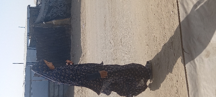 A young female, appearing to be a child or adolescent, is depicted in profile, facing right, standing on dusty, unpaved ground in Khan Yunis, Palestinian Territory. She is dressed in a full-length, dark blue garment with a floral pattern and a matching head covering. Her right hand is partially visible near her chest. A prominent shadow extends from her figure onto the sandy ground. In the background, multiple structures composed of corrugated metal sheets and dark tarpaulins, some displaying patterns, are visible under a bright, clear sky, indicating a temporary settlement or camp environment.