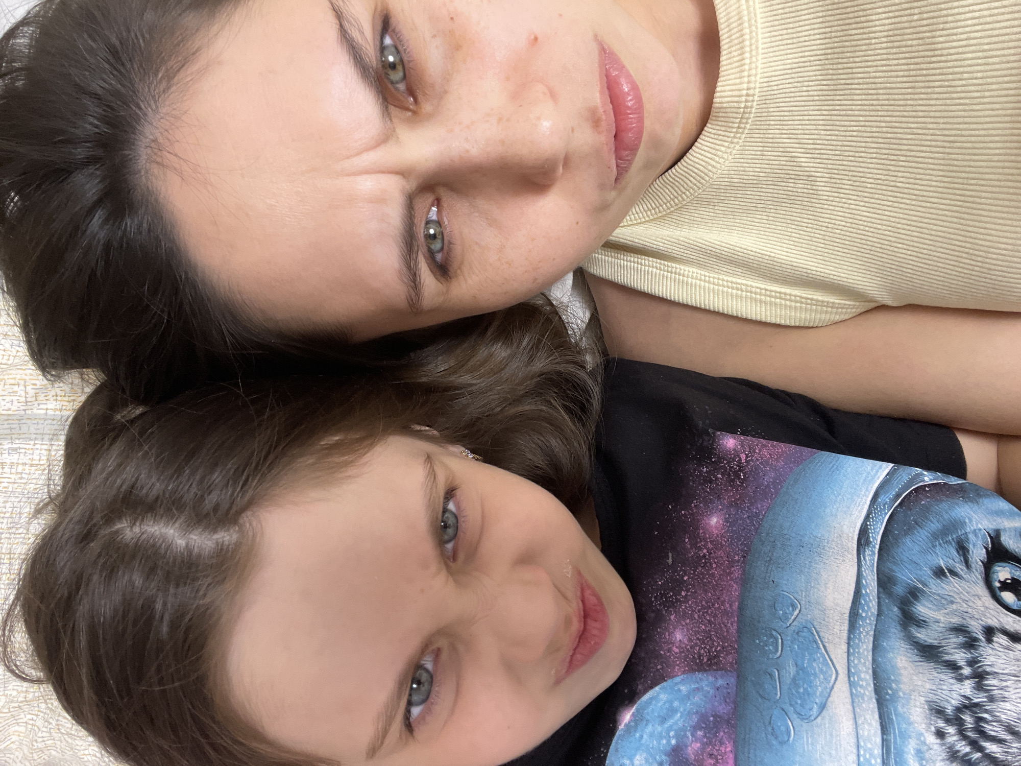 Two people, an adult and a child, are lying down next to each other. The adult has dark hair, green eyes, and is wearing a yellow shirt. The child has light brown hair, blue eyes, and is wearing a black t-shirt with a space-themed graphic. The background appears to be a light-colored fabric or surface. The location is not discernible from the image.
