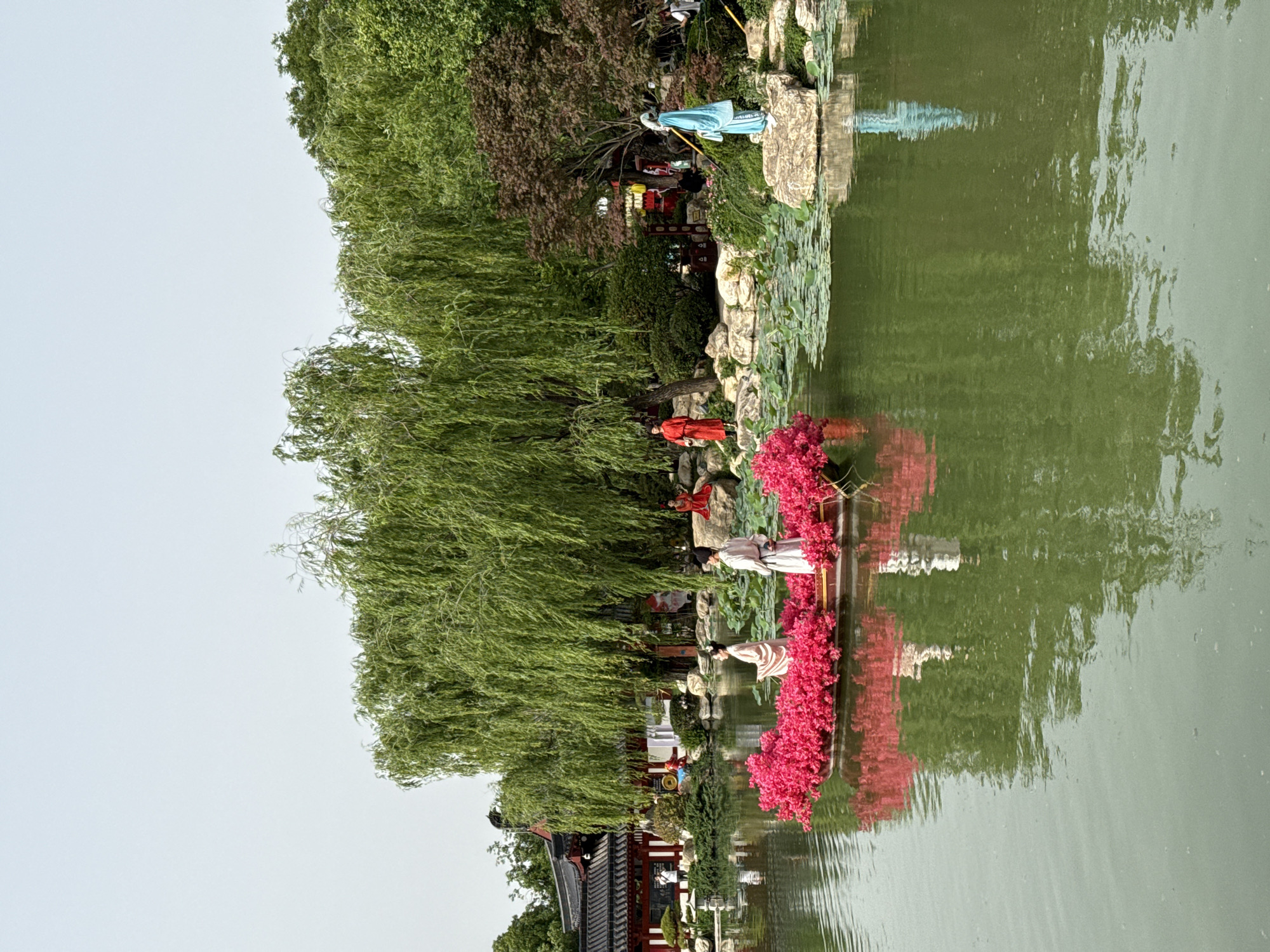 The image shows a garden scene, likely in the Lintong District of China, given the traditional architectural style and clothing of the figures. A body of water reflects the scene above.
Figures dressed in traditional Hanfu-style clothing are positioned near the water's edge. One figure, in white, stands next to a cluster of red flowers. Another figure is in red, located higher up on the terrain, close to a willow tree. Further along the water's edge, a figure in blue stands on a rock.
A traditional Chinese building with a dark roof and red columns is visible in the lower left. Behind the figures, various trees and greenery populate the landscape. The sky above is a pale blue or gray.