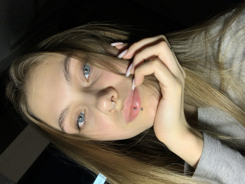 A young female subject is depicted in a close-up, head-and-shoulders portrait shot. She has light brown hair, blue eyes, and fair skin. Her facial features include a silver septum ring piercing and a curved barbell piercing on her lower lip. Her left hand, with long nails featuring light-colored polish, is positioned with her knuckles resting under her chin and fingers extending upwards towards her cheek. She is wearing a light grey, ribbed long-sleeved garment. The background is dark and out of focus, with a faint, indistinct light-colored rectangular object visible on the left side. No visual cues within the image indicate the specific location in the city of Фоки, Russia.