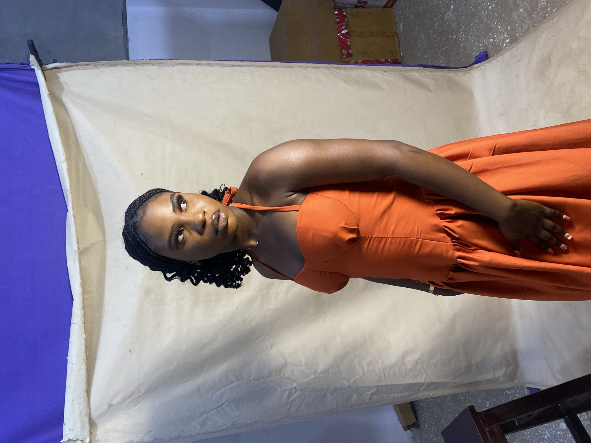 A woman stands in front of a backdrop consisting of off-white paper, supported by a purple sheet on one side. Above the backdrop are boxes. She is wearing an orange dress. The location appears to be a studio or temporary set-up, possibly in the city of Jos, Nigeria, although this is not definitively evident from the image. She is looking up and to her left.