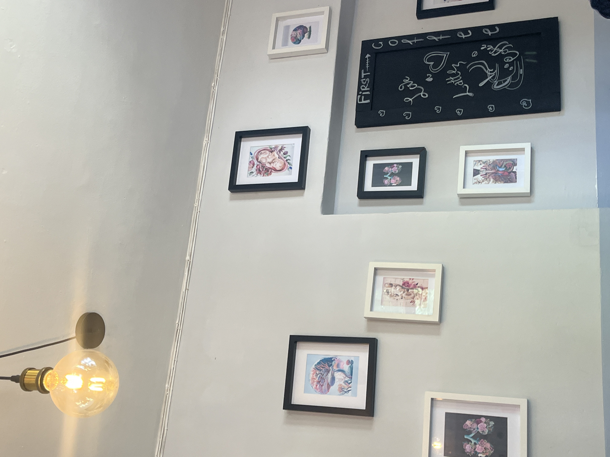 A wall with a collection of framed artwork is seen in the image. The wall is painted a light gray and has a few framed pictures of varying sizes and styles. Some of the frames are black, and some are white. The artwork includes images of flowers, human anatomy, and a chalkboard with writing. The bulb of a lamp is partially visible on the left side of the frame, near the bottom. The image has a sense of coziness and artistic display. The location appears to be indoors, likely in Plovdiv, Bulgaria.