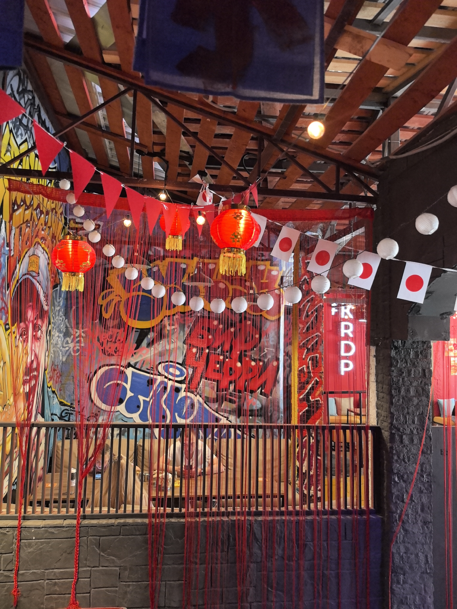 The image shows a dimly lit interior space decorated with Japanese cultural elements. Red lanterns and strings of white paper lanterns hang from the wooden ceiling beams. Several small Japanese flags are strung between the lanterns. The walls are covered in vibrant graffiti art. A black metal railing with vertical slats is in the foreground, with red strings hanging down in front of it. Beyond the railing, people can be seen sitting at tables. The overall atmosphere appears festive, possibly suggesting a restaurant or a bar. The location is identified as the city of Perm, Russia.