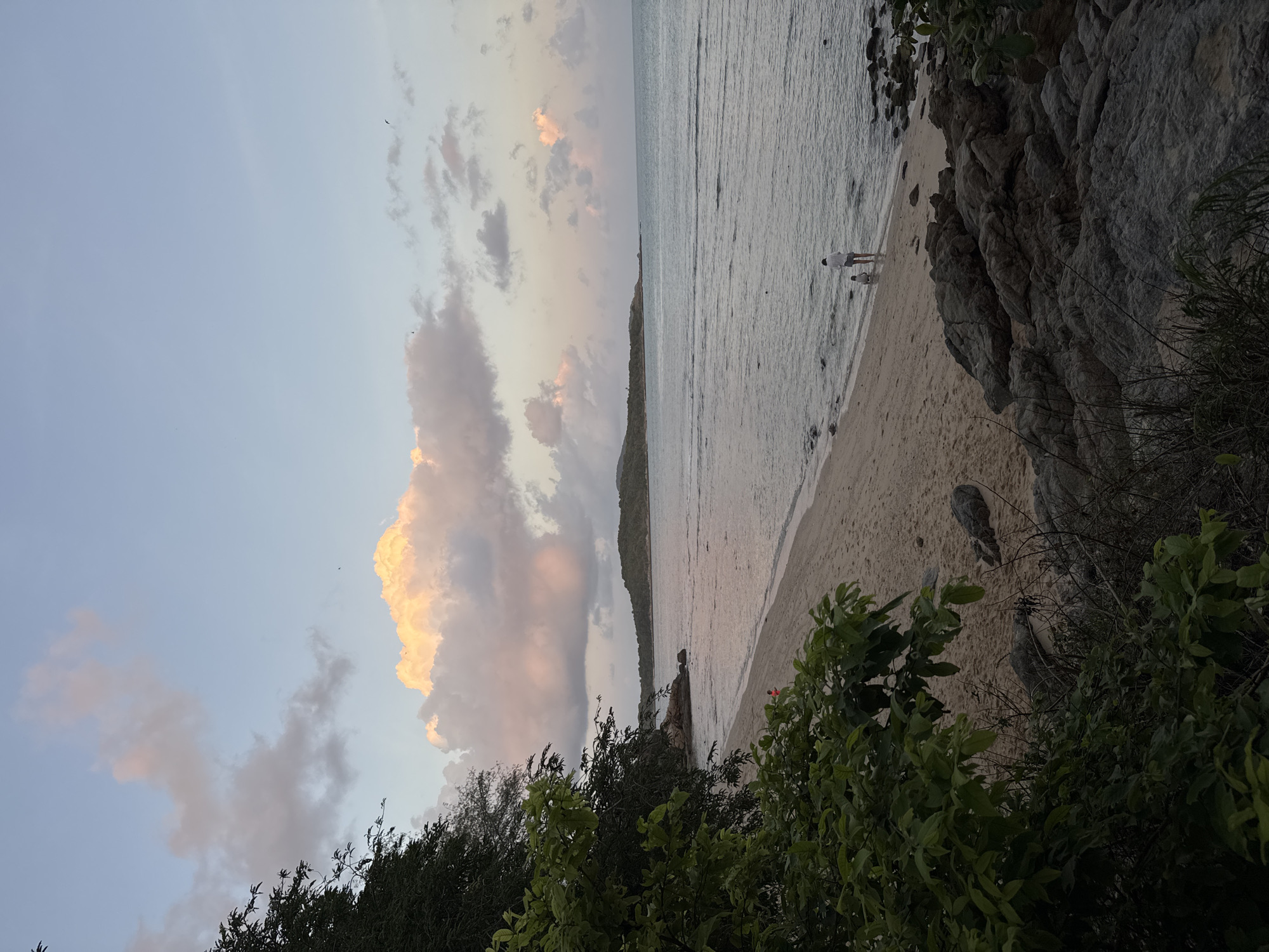 A full shot captures a serene beach scene at Rawai, Thailand, during sunset. The sky, a canvas of soft blues and pinks, is adorned with scattered clouds, some illuminated with a warm, peachy glow. The ocean, calm and reflective, gently laps at the sandy shore. Two figures, a child and an adult, stand near the water's edge, their silhouettes dark against the pastel hues. Lush green foliage frames the left and right sides of the image, hinting at the tropical environment. In the distance, a lush, green island rises from the sea, its contours softened by the evening light. Rocks, scattered along the shoreline, add a touch of ruggedness to the otherwise tranquil landscape.