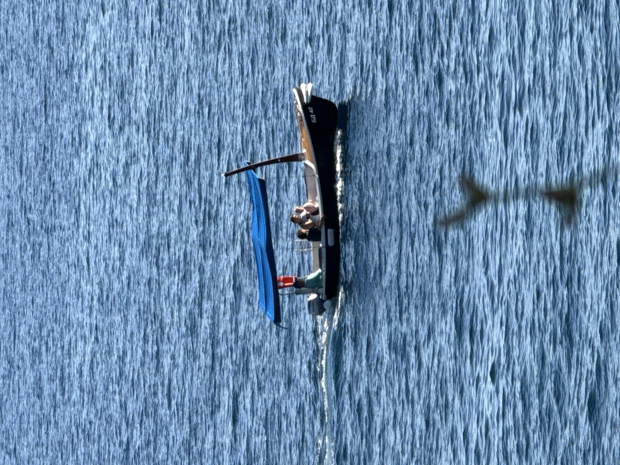A small motorboat with a blue canopy is moving through calm blue water. There are at least four people on board, two in the front and two in the back. The boat has a number, "ЕА 42", written on its side. The scene is set against a backdrop of rippling water, suggesting it is either the sea or a large lake. The bright sunlight indicates it is daytime. The overall impression is one of a leisurely boat trip on a sunny day. The location is the coast of Kamenovo, Montenegro.