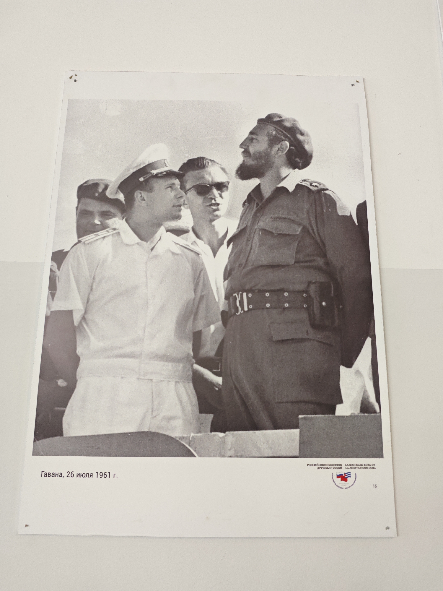 The image is a black and white photograph pinned to a wall. It depicts a group of men, most prominently featuring two individuals in the foreground. The man on the left is wearing a light-colored naval uniform with a peaked cap. The man on the right wears a dark uniform with a beret, belt, and holster. Both men are looking in the same direction.
The text at the bottom reads "Гавана, 26 июля 1961 г.," which translates to "Havana, July 26, 1961." To the right of the text is an emblem with text and a flag-like logo. Next to that emblem is the number 15.