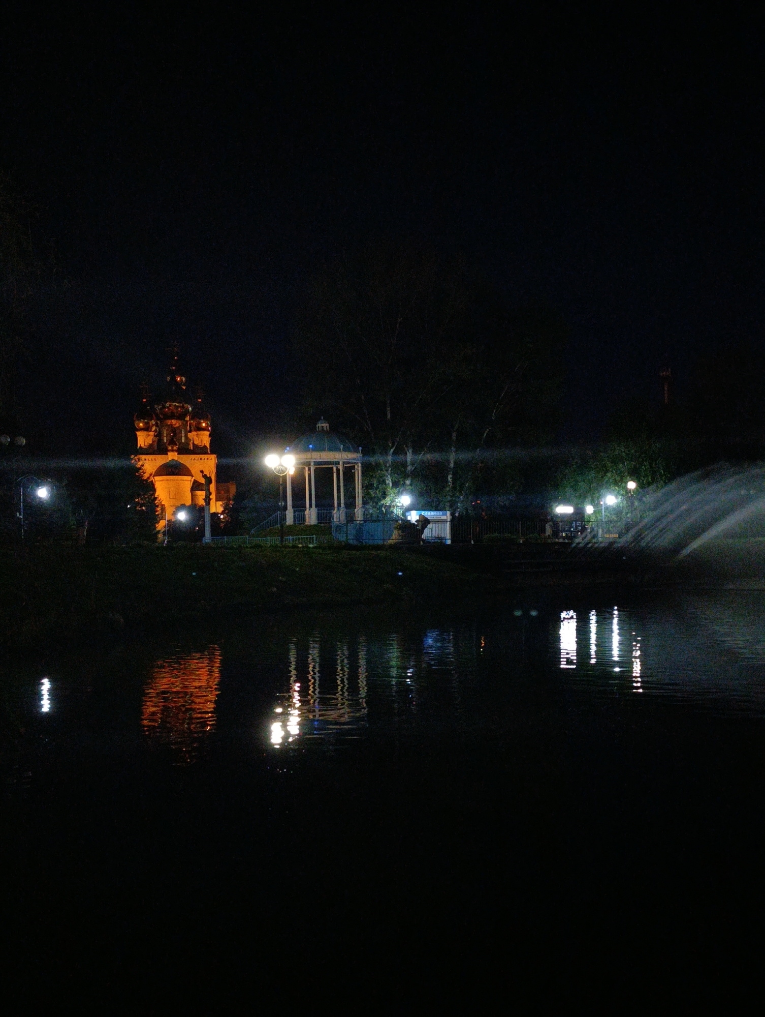 At night, a water body reflects illuminated structures along its bank. On the left, a tall, orange-lit building with multiple domes is visible, appearing to be a church. To its right, a white, octagonal gazebo with a domed roof is lit from within. Several streetlights illuminate the area. Further to the right, a fountain sprays water, creating a mist. Trees and foliage are present on the bank, some of which are illuminated by the lights. The scene is dark, with the lights creating bright spots and reflections on the water. This image was captured in Abakan, Russia, likely in a park or public area featuring these landmarks.
