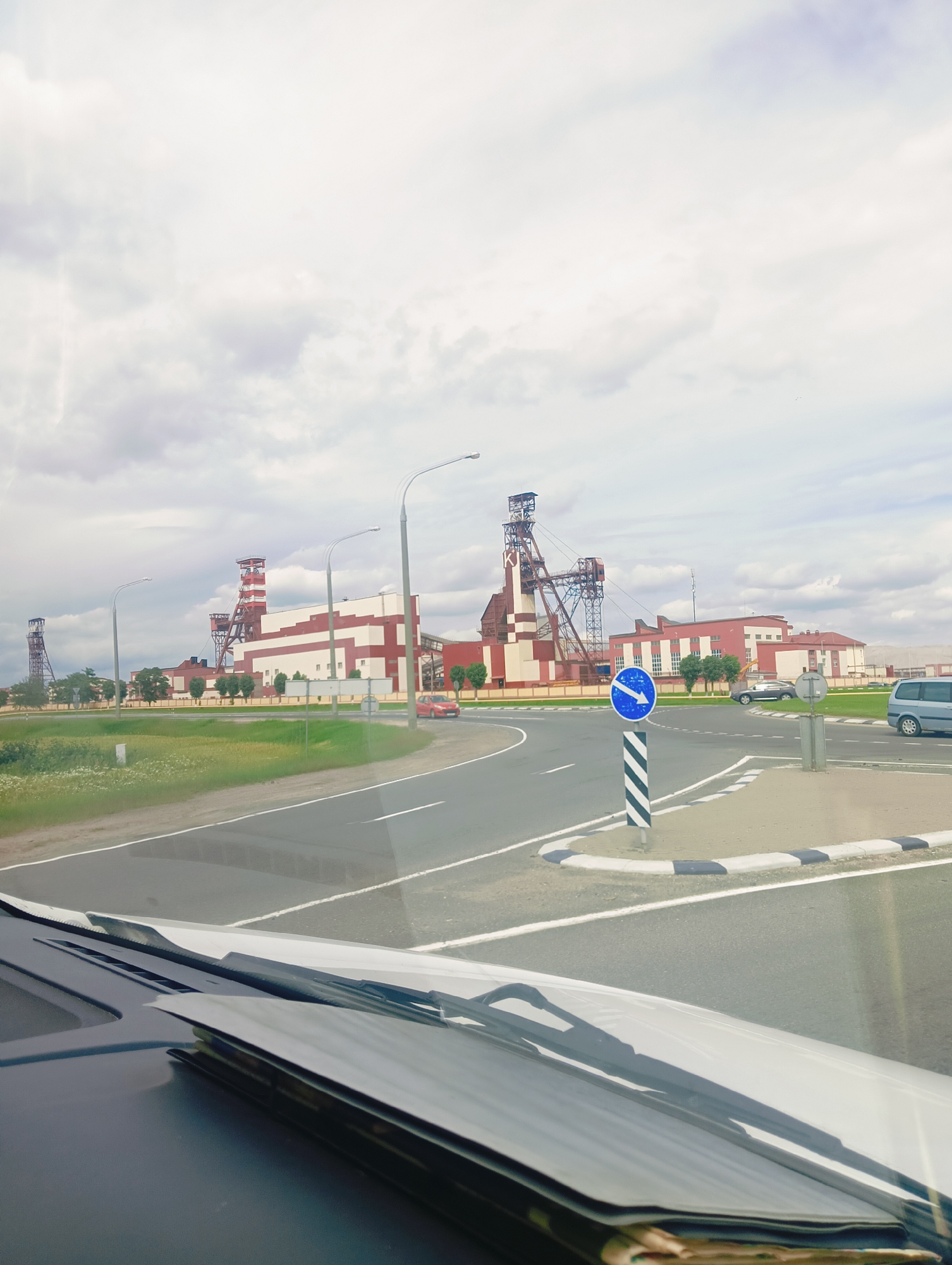 The image depicts a view from inside a vehicle, looking out at a large industrial complex under a cloudy sky. The complex features several tall, red and white structures, likely mine shafts or processing facilities, with distinctive lattice towers. In the foreground, a road curves to the right, with a traffic sign indicating a mandatory direction to the left. A black and white striped pole supports the blue circular sign with a white arrow.
The scene appears to be an operational industrial area, possibly a mining site given the prominent structures. The road is asphalt with white lane markings, and a patch of green grass and some trees are visible on the side. Several vehicles are present, including a red car on the road, a dark-colored SUV, and a light blue van further in the distance. The presence of the vehicles suggests ongoing activity at the location.
The weather appears overcast, with a bright, diffused light that doesn't cast harsh shadows, suggesting it might be daytime, but not necessarily midday. The overall mood is neutral, focused on the industrial landscape and the immediate surroundings of the road. There is no visible text in the image, apart from the symbols on the traffic sign. The location context provided is Краснадворскі сельскі Савет, Belarus, which helps identify the setting as an industrial area within this region.