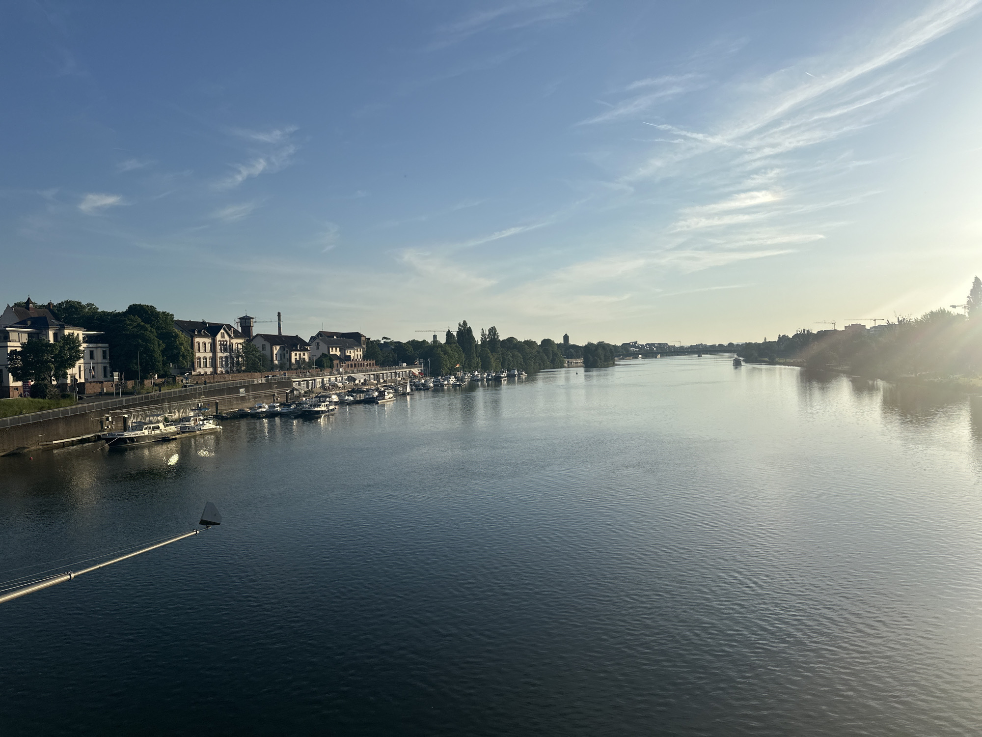 This is a wide shot of the Neckar River in Heidelberg, Germany on a clear day with a few wispy clouds and sunshine. The river takes up most of the image. On the left side is a shore lined with trees and a mix of white buildings with varying roof designs. There are several small boats docked along the shore. A tall, slender pole with a small, white, triangular flag at the top extends into the water from the lower left. On the right side of the river, there is land covered with trees and foliage, with the sun gleaming behind it. In the distance, there are glimpses of other structures and a crane. The river's surface is rippled.