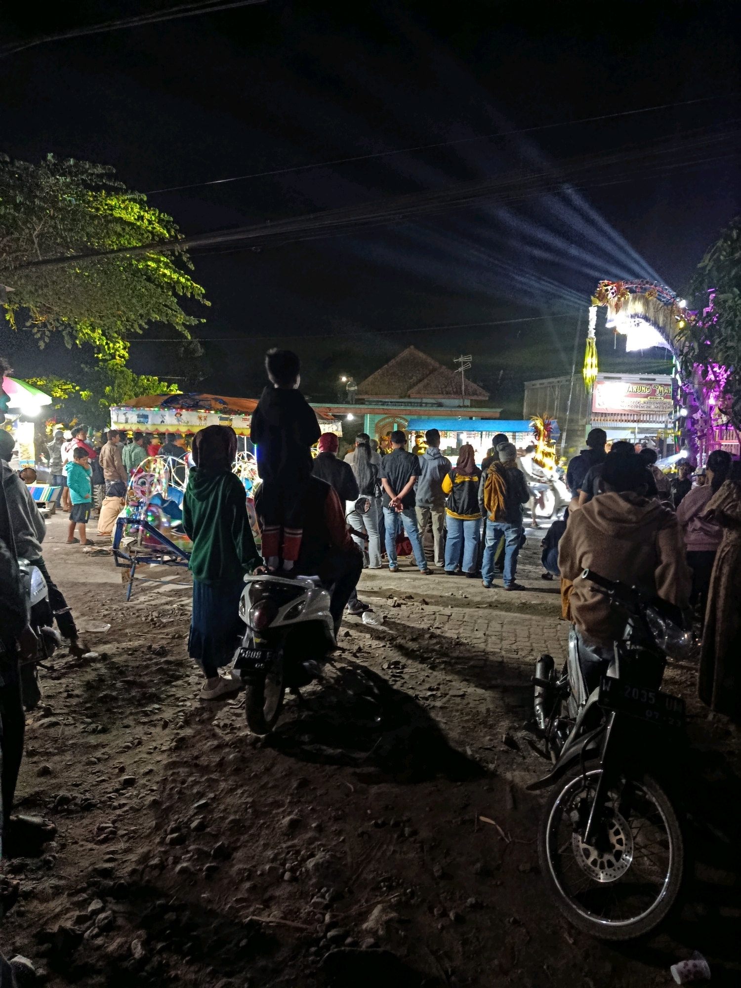 The image captures an outdoor scene at night in Talangsuko, Indonesia, likely a festive gathering or market. The foreground is dominated by rough, dusty ground with scattered rocks, illuminated by the artificial lights of the event. Two motorcycles are parked in the foreground, one closer and more detailed, showing a license plate that reads "W 2035 UM" and "07-23". In the middle ground, a crowd of people are visible, some standing and observing, others milling about. A child is seen perched on someone's shoulders in the center of the image, looking towards the right. Behind the people, a colorful carousel or amusement ride is present, illuminated with lights. Further in the background, there are structures that appear to be stalls or buildings, with more lights and decorations, including a prominent archway adorned with lights and what looks like a dragon motif. The overall atmosphere suggests a lively night event, possibly a fair or festival, taking place on a dirt or unpaved area. The sky is dark, indicating it is nighttime, with visible power lines and street lights.
