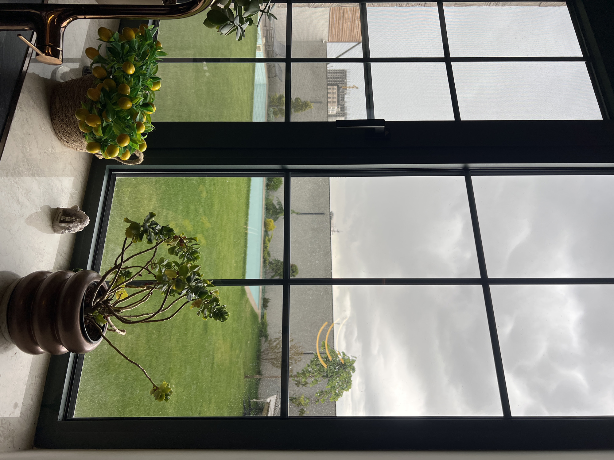 The image shows a view through a window, likely in a building in Yerevan, Armenia based on visible architectural styles. The window has a black frame and multiple panes. Outside, there is a well-maintained green lawn with a blue-edged pool. Beyond the lawn is a light-colored concrete wall. In the distance, a construction site with a crane and an apartment building can be seen. The sky is overcast with thick gray clouds.
Inside, on the window sill, there is a brown, ribbed pot with a sprawling plant. To the left, there is a small stone object and a straw basket containing a decorative lemon tree. A copper-colored faucet is also visible in the upper-left corner of the image.