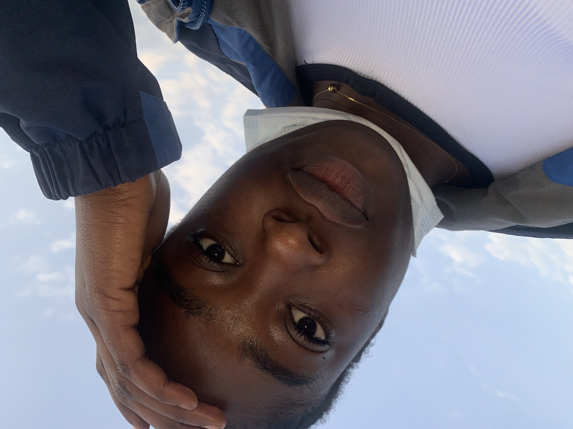 The image is a close-up, angled selfie of a young Black person outdoors. The subject is looking towards the camera with their left hand resting on their forehead. They are wearing a blue and gray jacket over a white ribbed tank top, and a blue mask is hanging around their neck. The background is a clear blue sky with scattered white clouds, suggesting daytime. The lighting indicates the sun is positioned to the subject's left, highlighting their skin. The image does not contain any information or visual cues that would allow for the identification of a specific location in Rafin Par, Nigeria.