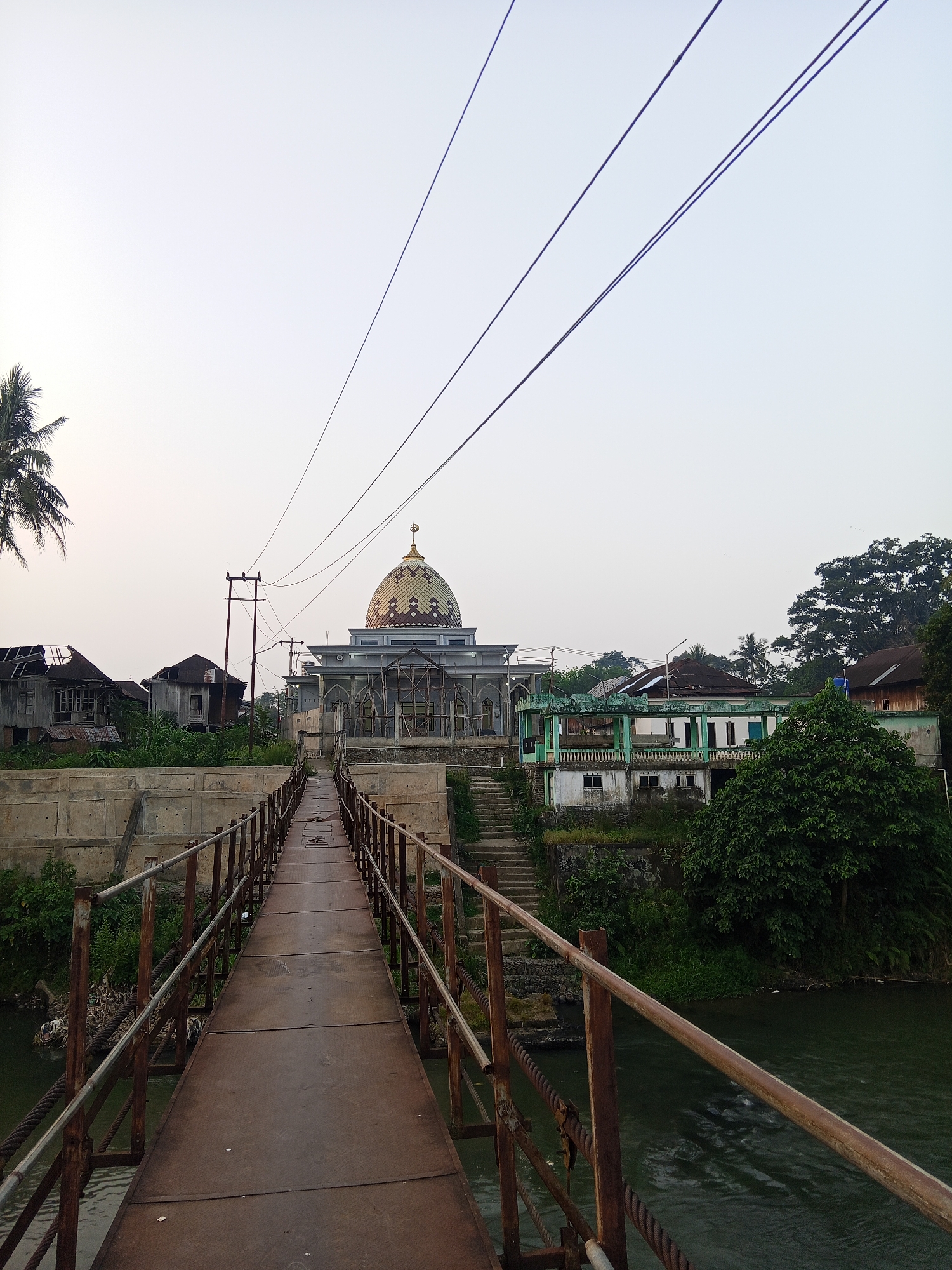 The image shows a pedestrian bridge constructed of rusted metal, leading towards a mosque with a golden dome in Lubuklinggau, Indonesia. The bridge spans a body of water, which appears to be a river, with visible currents. On the left side of the bridge, there are several dilapidated buildings with damaged roofs. On the right side, there is lush green vegetation and more buildings, including the mosque which is the focal point of the scene. The sky is overcast and bright, suggesting it is either early morning or late afternoon. Several electrical wires are visible strung across the sky. The overall scene depicts a somewhat neglected urban or semi-urban environment with a prominent religious structure.