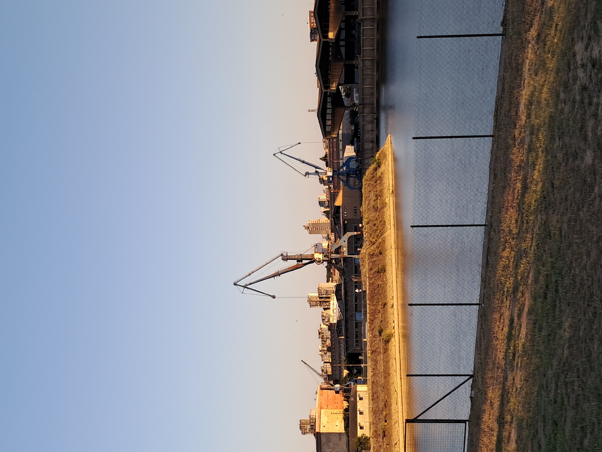 The image depicts a wide-angle, outdoor view of a port or industrial area along a body of water, likely a river or canal, in Belgrade, Serbia. The sky is clear and a pale blue, with the sun casting long shadows, suggesting it is either early morning or late afternoon. In the middle ground, several large cranes are prominent, indicating shipping or loading activities. These cranes are industrial in design, with their boom structures reaching high into the air. Behind the cranes, there are buildings that appear to be warehouses or industrial facilities, some with multiple stories and exhibiting a utilitarian architectural style. In the far background, a cityscape is visible, characterized by apartment buildings and other urban structures. The foreground features a grassy area with dry patches, leading to a chain-link fence that borders the water. The water itself appears calm, reflecting the light and the surrounding structures. The overall impression is one of an active, but possibly older, industrial waterfront.