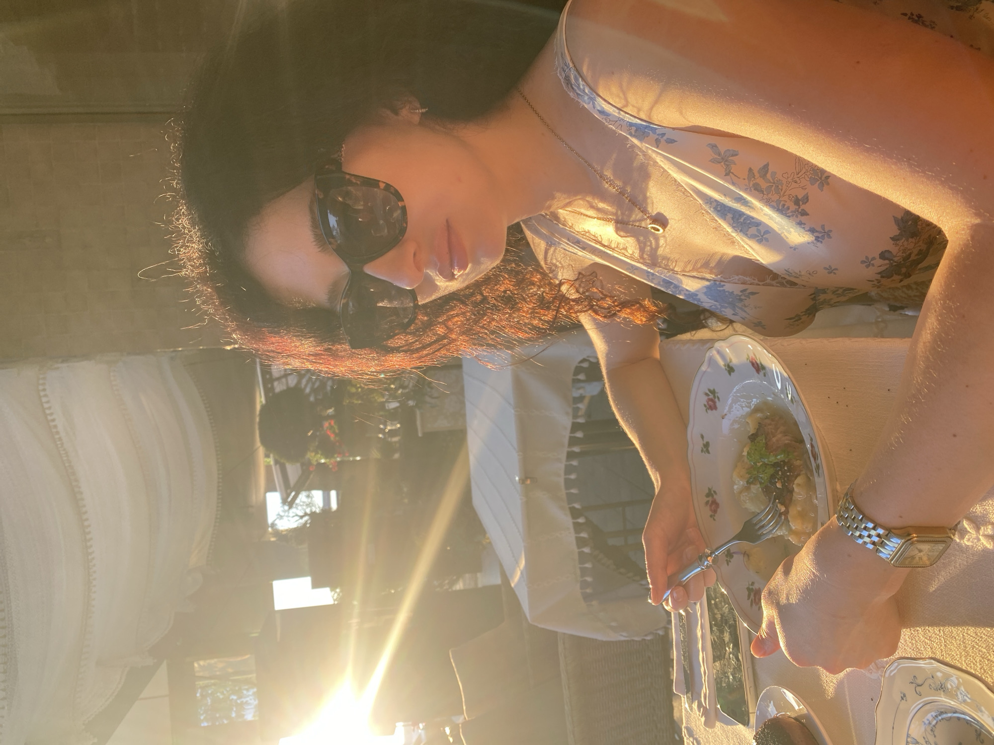 An adult female is seated at an outdoor or semi-outdoor dining establishment in Sochi, Russia, during the golden hour, indicated by direct, low-angle sunlight creating lens flare from the left. She has dark, curly hair with sun-illuminated red highlights and wears tortoise-shell sunglasses, a light-colored sleeveless garment with a blue botanical pattern, a gold necklace with a circular pendant, and a small earring in her right ear. On her left wrist is a rectangular gold-toned watch with a link band. She holds a fork in her right hand and a knife in her left, positioned over a plate containing what appears to be mashed potatoes with a meat patty and green garnish. The table is covered with a white tablecloth featuring a tasseled trim. To the left of her plate are a face-down smartphone, a small decorative plate with a blue floral pattern, and a dark round item on another white plate. Wicker chairs with cushions are visible in the background, along with a white fabric canopy with tassels overhead. Distant structures and foliage, including a small manicured tree, are also visible under the golden light.