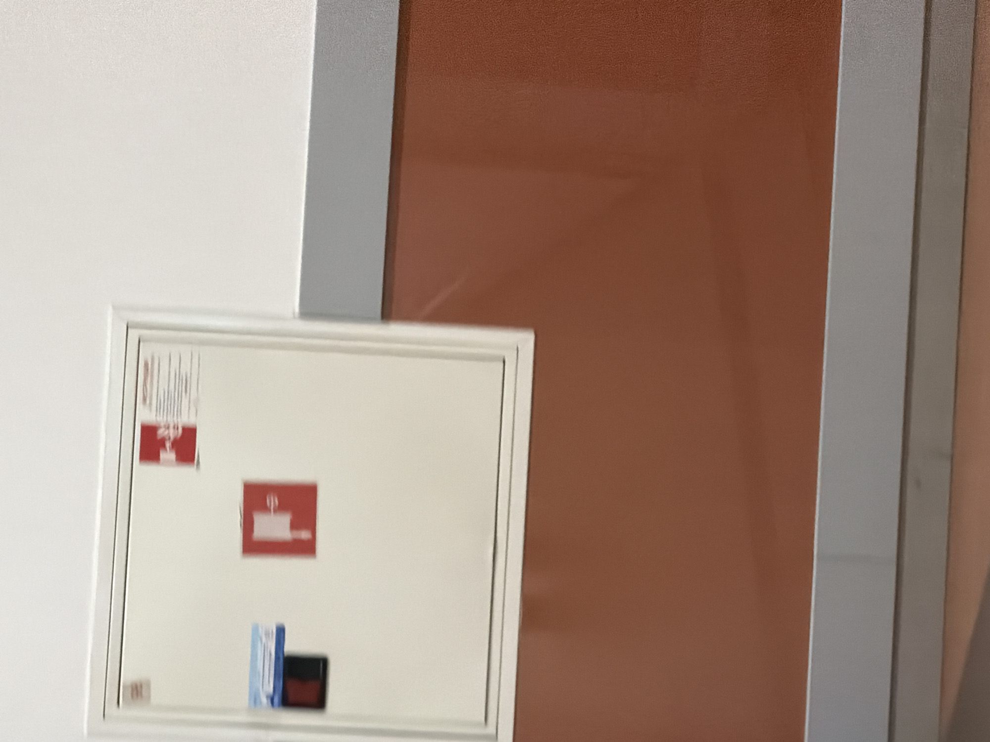 This is a medium-shot, eye-level, indoor photograph showing a white cabinet mounted on a wall. The cabinet is slightly ajar and has several red and blue stickers on its surface. One red sticker, on the top right, appears to be a fire safety symbol with text. Below that, on the left side of the cabinet, are two blue stickers, one with a dark blue strip. A red sticker with a white graphic of a tool is located in the center of the cabinet.
The wall behind the cabinet is painted white. Below the cabinet, the wall transitions to a reddish-brown color, separated by a thin gray stripe. The bottom of the image shows the floor, which appears to be a light brown, possibly tiled surface. The overall environment suggests an interior space, likely a public building or facility, such as a school, hospital, or office. The image does not provide any specific geographical clues that would allow for pinpointing its location within Sofia, Bulgaria.