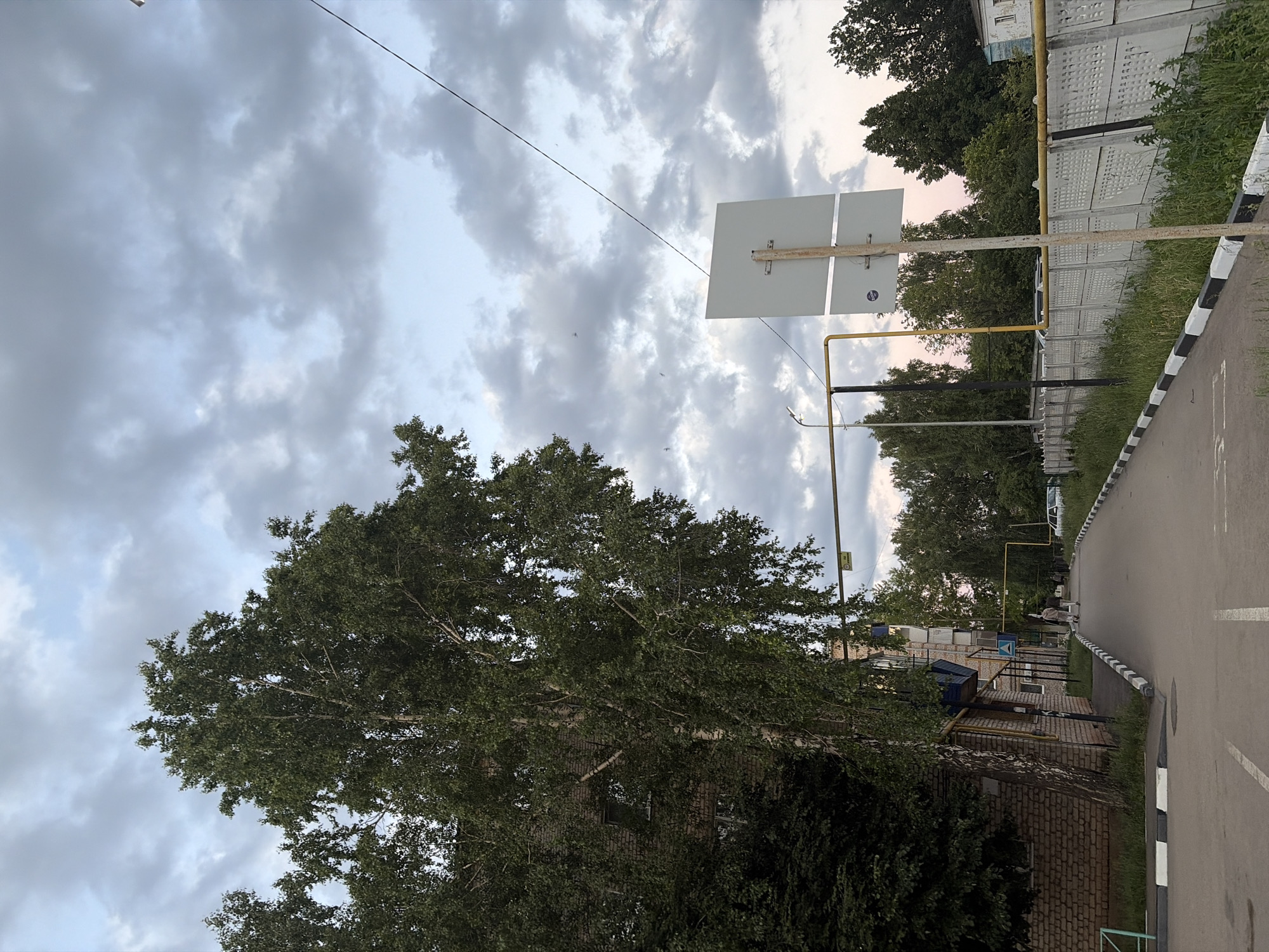The image shows a street in the city of Ilchimbetovo, Russia, on a cloudy day. A tall birch tree stands to the left, with its branches extending over a brick building. A paved road runs through the center of the image, bordered by a concrete fence and a sidewalk. There are a few people walking on the sidewalk and a car parked in the distance. A white sign on a pole is visible on the right side of the road. The sky is filled with grey clouds, suggesting an overcast day.