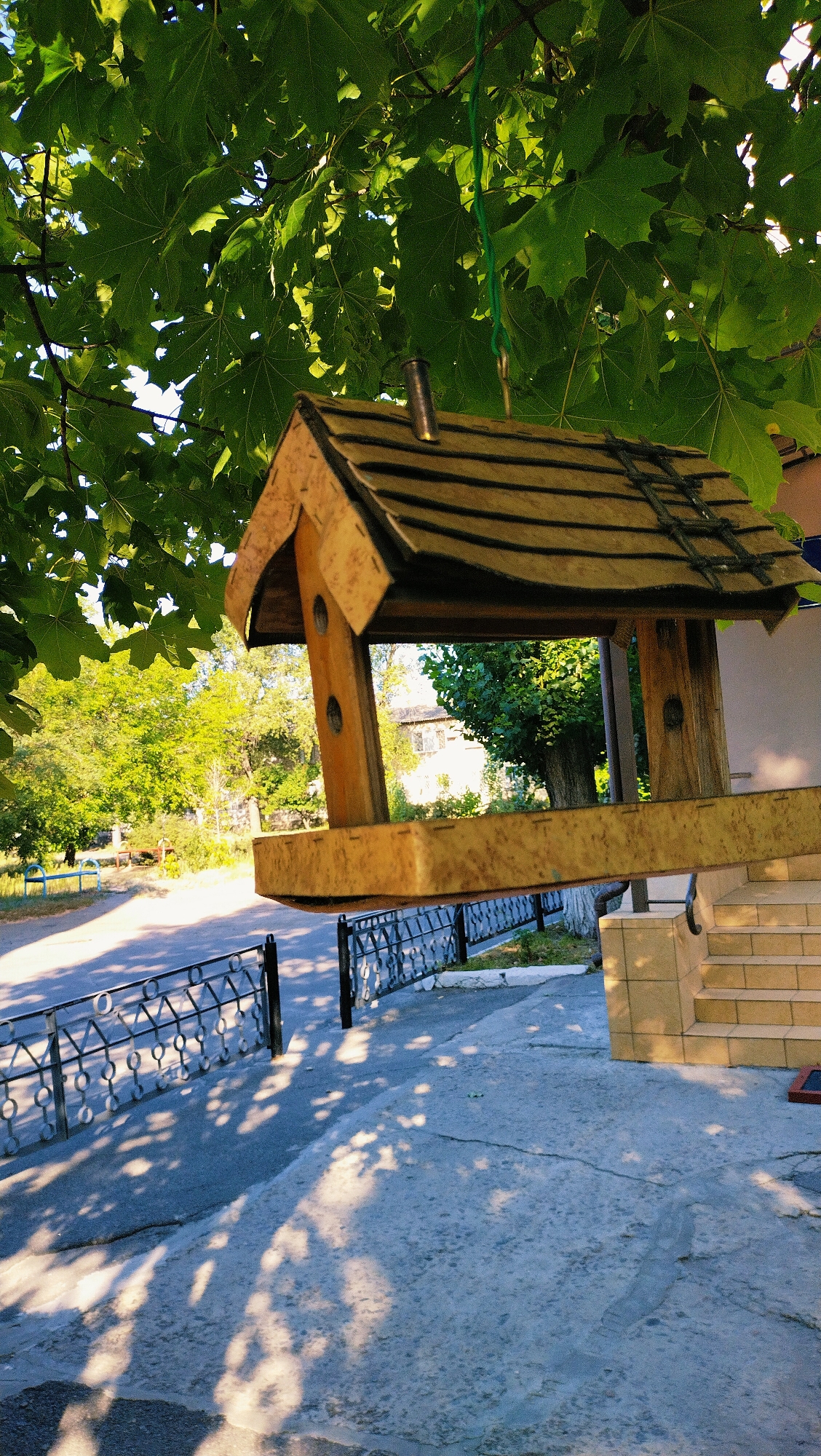 A wooden bird feeder hangs from a tree with green leaves. The feeder is light brown and has a slanted roof. In the background, there is a paved area with a black metal fence and a set of stairs leading to a building. The scene appears to be in a residential area, possibly a park or a courtyard. The image is taken from a low angle, looking up at the bird feeder. The lighting suggests it is daytime, with sunlight casting shadows on the ground. The location is the city of Horishni Plavni, Ukraine. There are no people visible in the image.