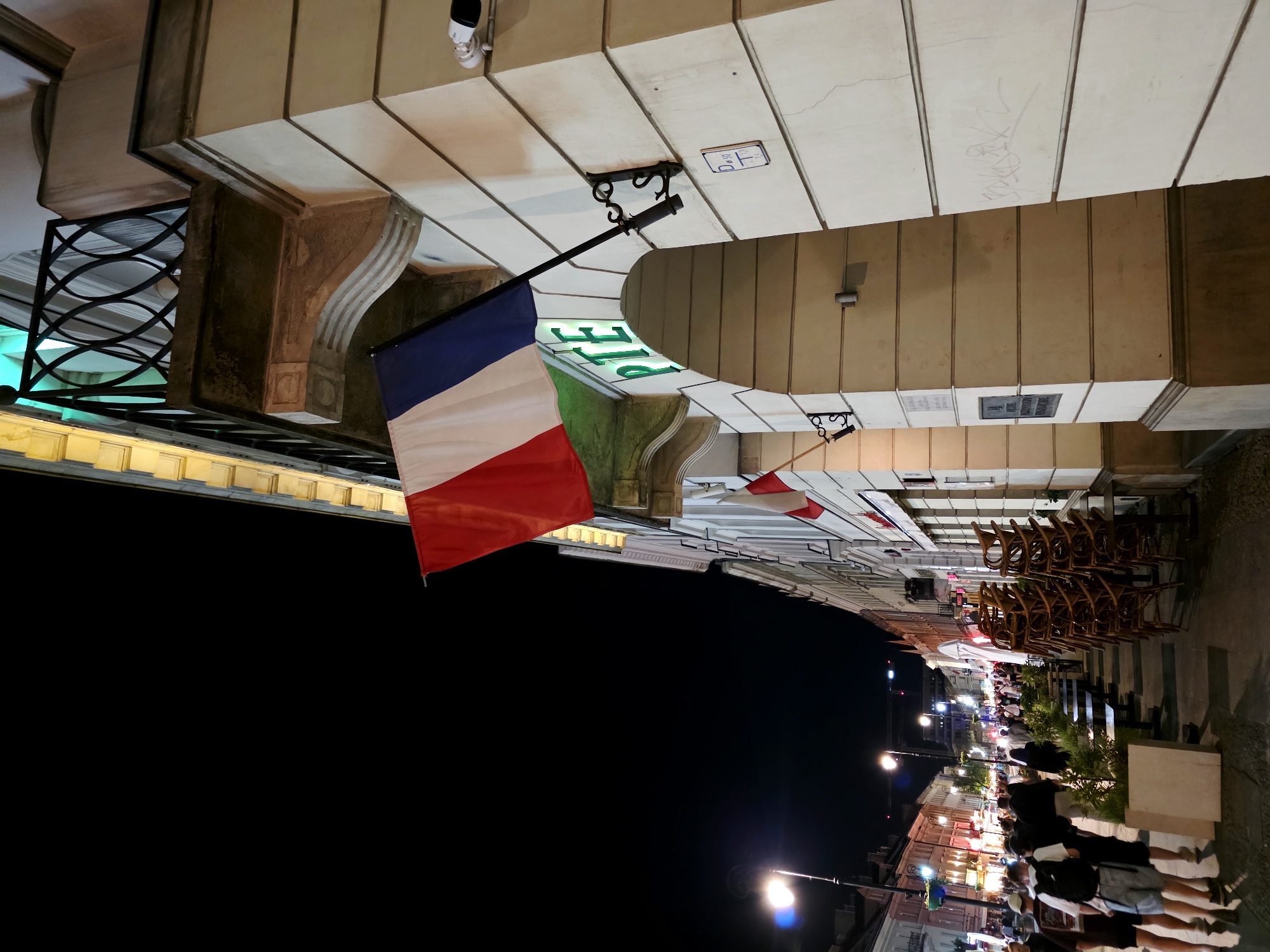 The image captures a street scene at night in Warsaw, Poland, specifically on Krakowskie Przedmieście street. A French flag is prominently displayed, attached to the facade of a building. The building's architecture features a balcony with an ornate railing and decorative stone elements. Adjacent to the balcony, the letters "PTE" are illuminated in green, suggesting a business or institution. Below the "PTE" sign is an arched doorway. Further down the street, other buildings with illuminated windows and signage are visible, as well as streetlights illuminating the pedestrian walkway. A line of stacked chairs and tables indicates an outdoor seating area for a restaurant or cafe, likely closed for the night. People are walking along the sidewalk, dressed casually, indicating a lively evening atmosphere. The overall impression is of a well-lit, urban street at night, with architectural features and visible signage contributing to its distinct character.