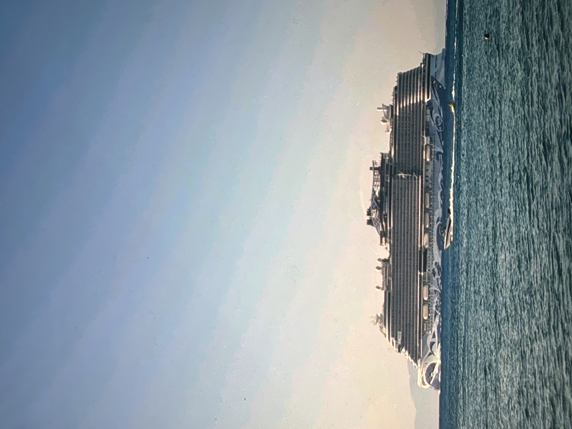 A large cruise ship sails across a serene, blue-green ocean under a clear, pale blue sky during daylight hours. The image is oriented to show the ship spanning horizontally across the right side of the frame, with the expansive sky filling the left.
The multi-decked vessel features a white and grey superstructure with numerous balconies visible along its sides. A distinctive hull art adorns the ship's side, characterized by intricate white and blue swirling, abstract patterns, possibly depicting waves or ice formations. This design is typical of modern cruise liners, such as those in the Norwegian Cruise Line fleet.
The ocean displays gentle ripples and texture on its surface, reflecting the light. A small, dark, spherical object, possibly a buoy, floats in the water towards the upper right section of the ocean. The sky is uniformly light blue, fading slightly towards the horizon, devoid of clouds, indicating fair weather. The overall lighting suggests a bright, calm day, possibly morning or late afternoon, with soft illumination across the scene. No people are visible, and there is no discernible text on the ship or elsewhere. The setting appears to be open water with no land in sight.
