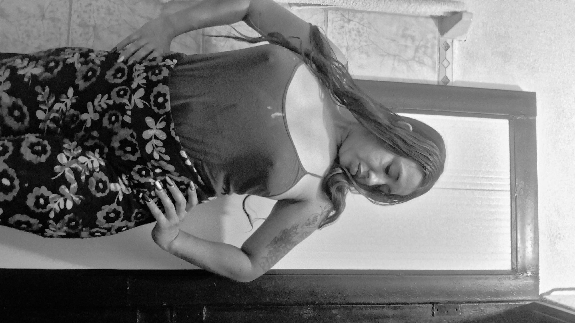 A grayscale, waist-up, medium shot shows a woman with long, dark, wavy hair and a tattoo on her right arm standing with her hands on her hips. She is looking down and to her right. She is wearing a dark, thin-strapped tank top and a dark skirt with a floral pattern. The background consists of a tiled wall on the left, a mirror, and a dark doorway on the right. The lighting is dim, and the overall impression is that of a candid, personal photograph taken in a bathroom.