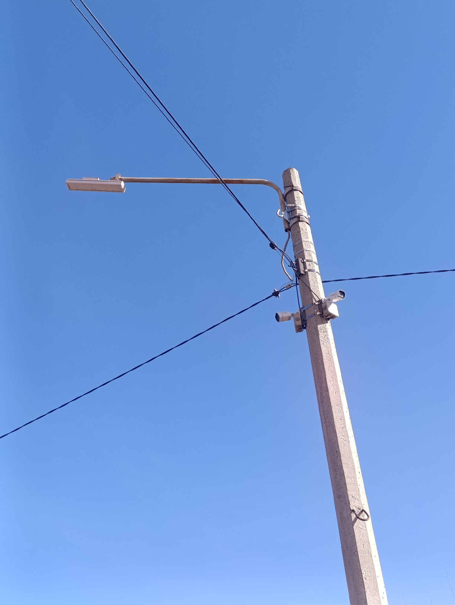 A low-angle shot depicts a concrete utility pole extending upward against a clear, cloudless blue sky. At the top of the pole is a street light fixture extending horizontally to the left. Below the light fixture, two security cameras are mounted to the pole with metal brackets. Electrical wires are strung across the top of the pole and across the sky in the background. A curved metal fitting is visible further down the pole. Based on the objects and architecture, this scene is likely located in Krasnoyarsk, Russia.