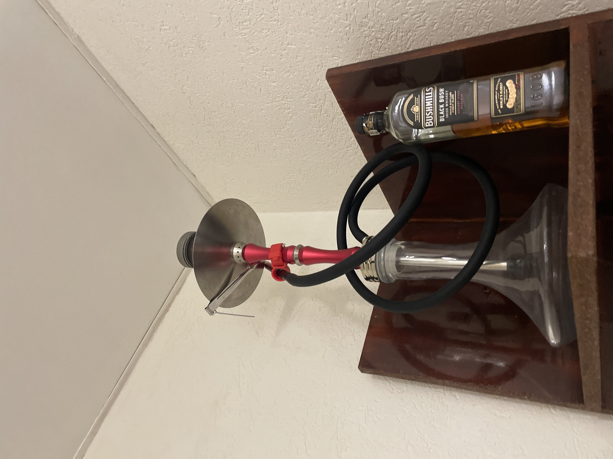 A hookah is positioned next to a bottle of Bushmills Black Bush Irish Whiskey on a dark wood shelf mounted on a white wall. The hookah has a silver bowl, a red stem, a clear glass base, and a black hose. The wall is likely indoors. The city of Sofia, Bulgaria is not discernible.