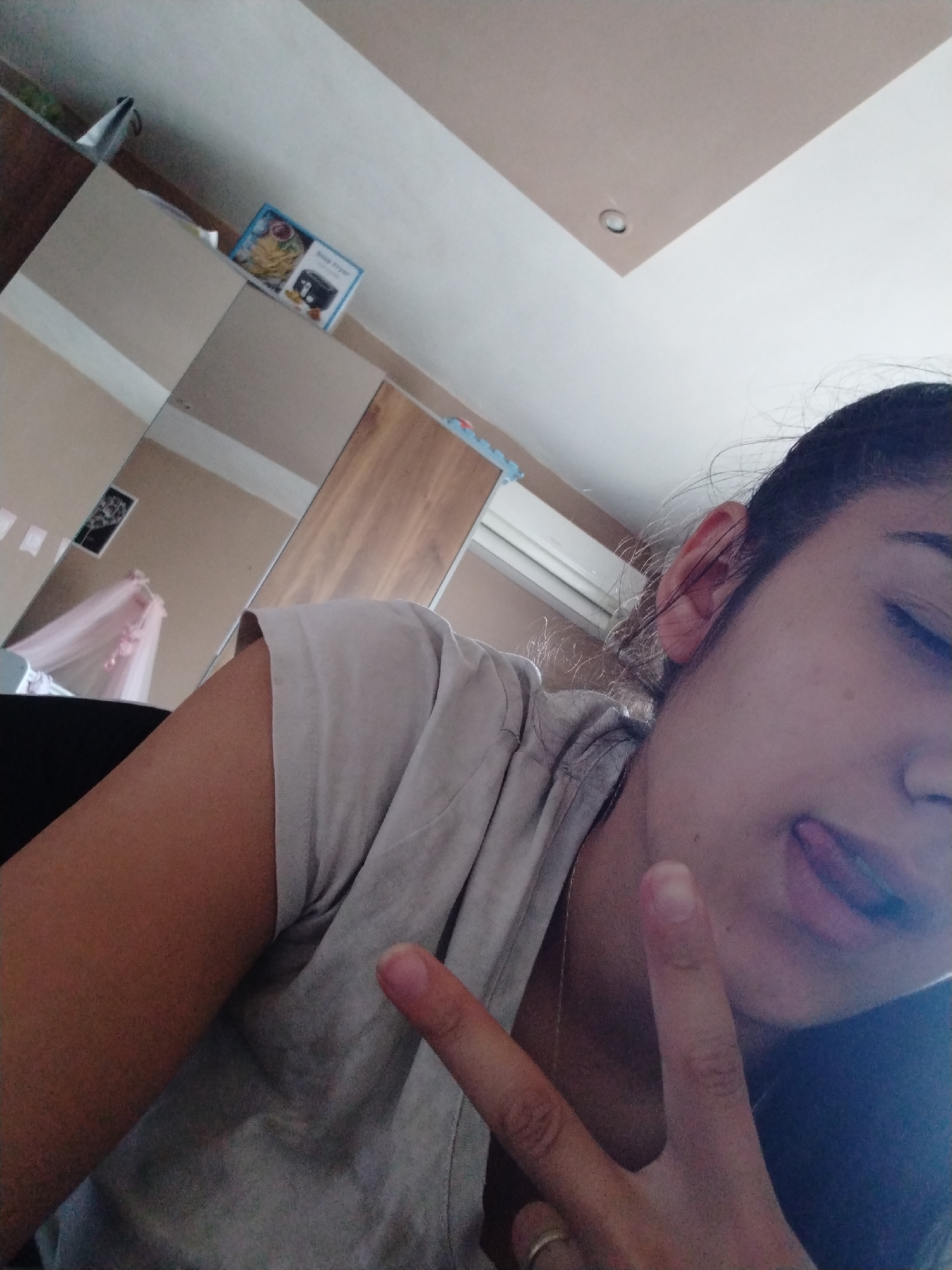 A selfie taken at an upward angle shows a person with dark hair, pale skin, and closed eyes sticking their tongue out slightly. They are wearing a light-colored t-shirt. The person is making a peace sign with their hand. In the background, there is furniture including what appears to be a mirrored wardrobe. There is also a box with an image of food on top of the wardrobe. The location is Unknown, Unknown.