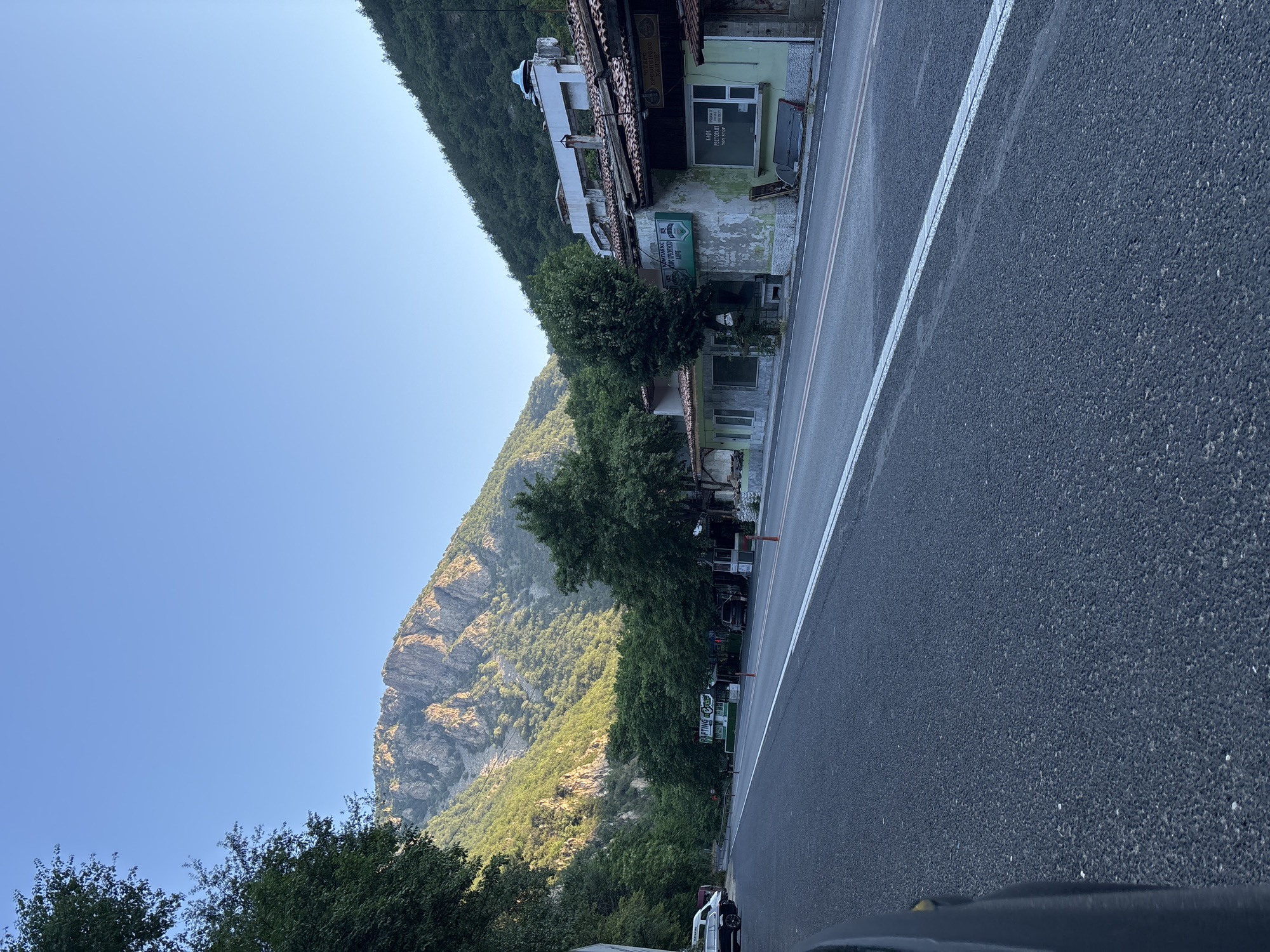 The image depicts a roadside scene in the city of Stara Kresna, Bulgaria. A paved road with white markings runs through the frame, flanked by a row of buildings and lush greenery. In the background, a majestic mountain range rises, its rugged slopes illuminated by the soft glow of sunlight. Trees densely cover the lower parts of the mountains, creating a stark contrast with the rocky peaks.
On the right side of the road, a collection of buildings appears to house businesses, indicated by signs. One building, painted in a faded green, displays a sign that reads "RAfting". Another sign above a storefront reads "КАФЕ РЕСТОРАНТ" (Cafe Restaurant). The buildings exhibit signs of age, with some appearing weathered and in need of repair. The architecture suggests a rustic, perhaps traditional style, with tiled roofs and stucco facades.
Several vehicles are visible. A white van and a dark-colored car are parked on the left side of the road. On the right side, near the buildings, a dark SUV is parked. Orange traffic cones are placed along the road. The overall atmosphere is one of a rural or semi-urban setting, characterized by a blend of natural beauty and small-scale commercial activity. The sky is clear and blue, suggesting fair weather.