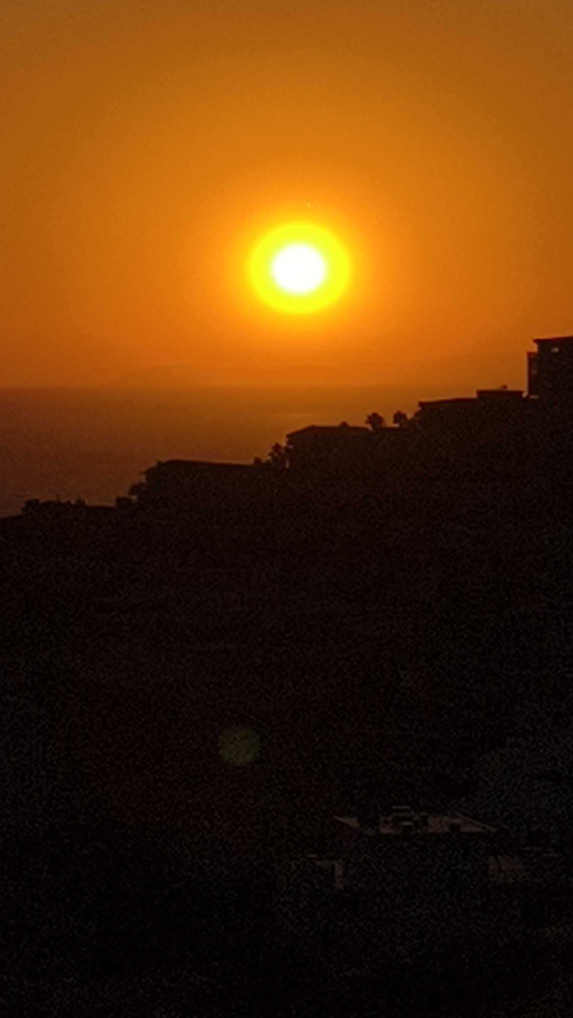 The image shows a sunset over Kuşadası, Turkey. The sun is a bright orange orb in the sky, with a halo of yellow light around it. The sky is a gradient of orange and yellow. In the foreground, there is a silhouette of buildings on a hillside. The buildings appear to be houses or villas. The hillside is dark and appears to be covered in trees. The sea is visible in the background, and it is a deep blue color. The overall impression of the image is one of peace and tranquility.