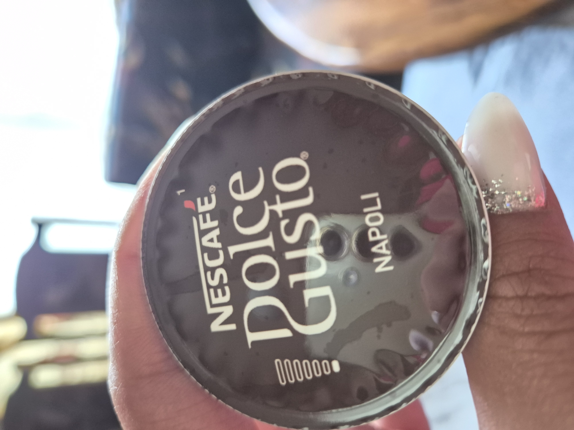 A human hand, characterized by medium-dark skin tone, holds a Nescafé Dolce Gusto "Napoli" coffee capsule. The hand's thumb and forefinger are visible, gripping the circular, dark-brown capsule. A manicured fingernail with light-colored polish and glitter accents is visible on what appears to be the ring finger. The capsule displays "NESCAFE" at the top, followed by "Dolce Gusto" in a larger script. Below this, "NAPOLI" identifies the coffee blend. A vertical sequence of eight small circles, with the top one filled, is present on the left side of the capsule, likely indicating intensity. A small red marking with the number "1" is located near the "NESCAFE" logo. Three small, circular indentations are visible near the center of the capsule's surface. The background is significantly blurred, indicating a shallow depth of field, and shows indistinct light and dark areas consistent with an indoor setting. No specific geographical markers or contextual elements are present in the image to determine a location in the city of Iskrets, Bulgaria.