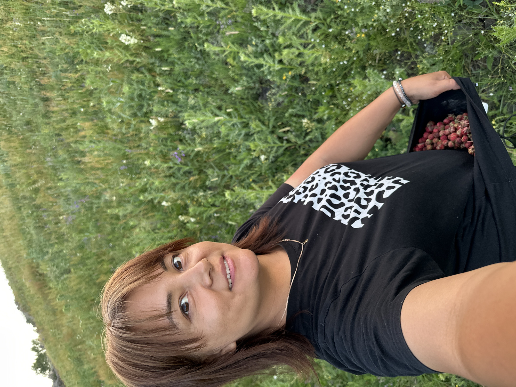 A woman is taking a selfie in a field of green grass and wildflowers. She is wearing a black t-shirt with a white leopard print design on the front and is holding a black bag filled with red berries. Her hair is brown and she has brown eyes. The background is a hillside covered in lush green vegetation.
