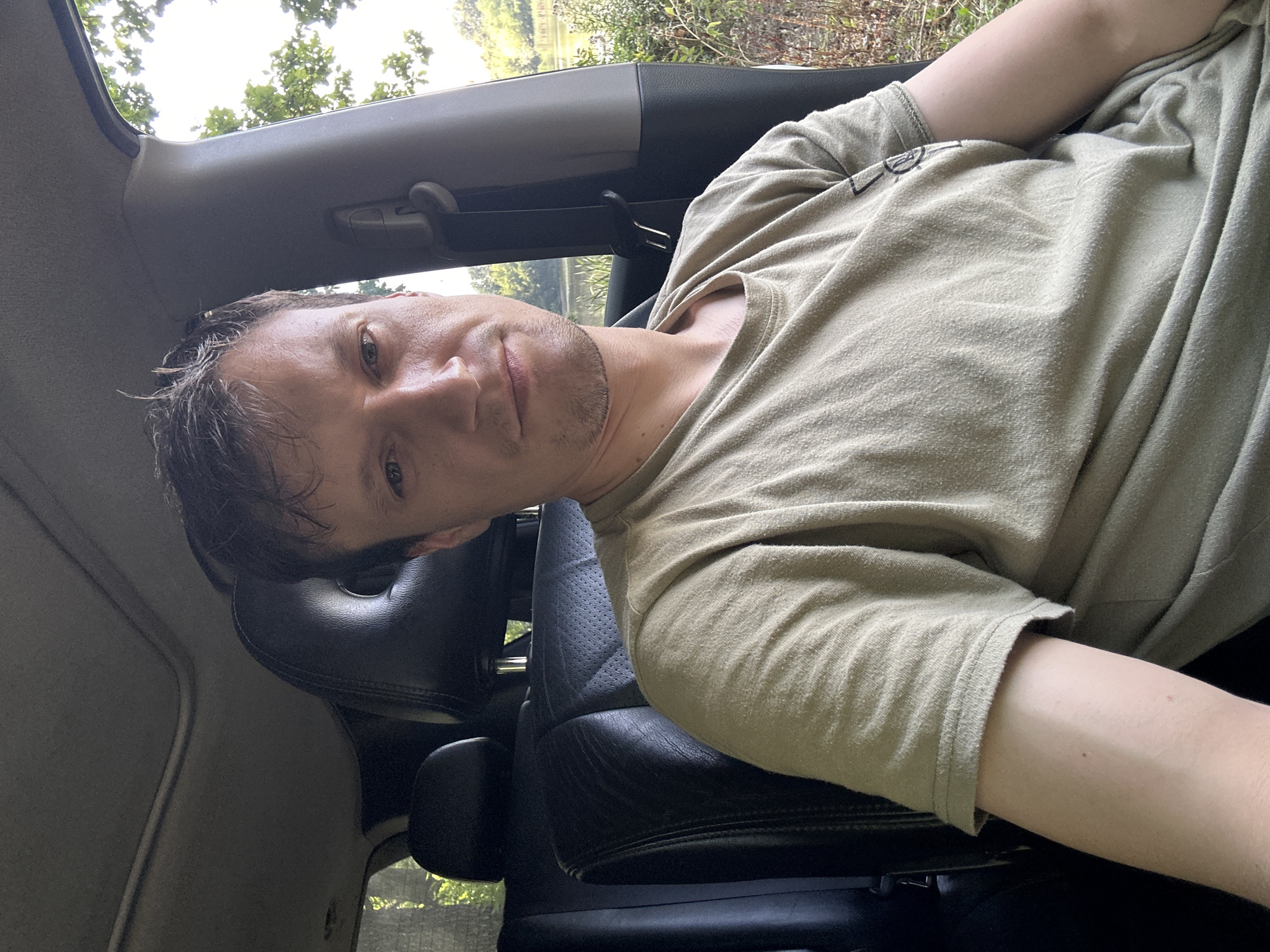 A medium shot shows a man in his 30s, with short brown hair and a light beard, sitting in the driver's seat of a car. He is wearing a light green t-shirt. The interior of the car is visible, with black leather seats and a sunroof. Outside the car, trees and a body of water can be seen, suggesting a rural or park-like setting. The man is looking directly at the camera with a neutral expression. The image appears to be a selfie taken during the daytime. There is no indication of the specific location within Makiv, Ukraine.