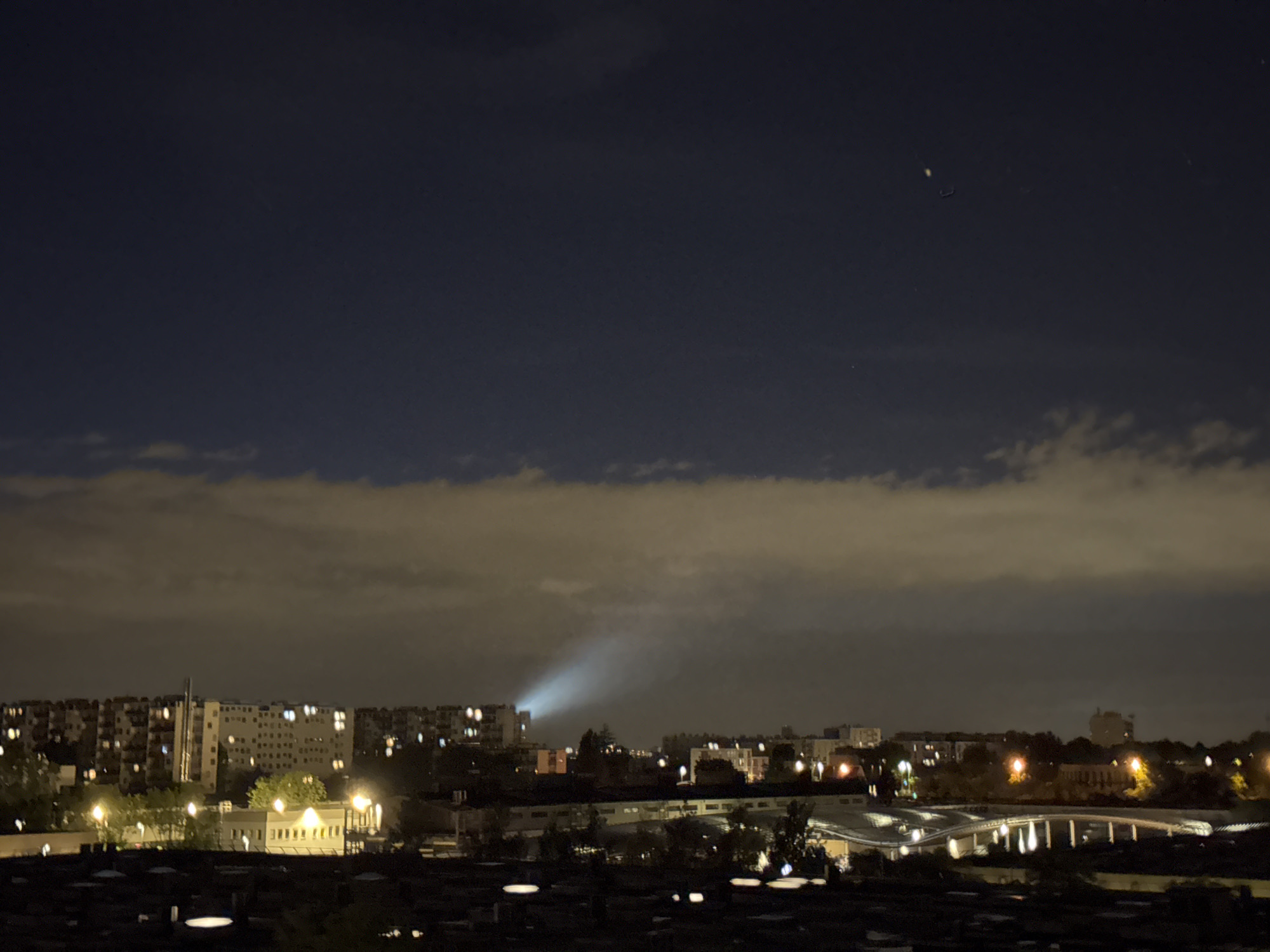 This is a long-exposure, night-time, elevated view of a city skyline. The sky is dark blue with scattered clouds, some of which are illuminated from below by city lights. A powerful beam of light emanates from one of the buildings in the distance, cutting through the clouds. In the foreground, the dark silhouettes of rooftops and trees are visible. The middle ground features a collection of buildings with lights on in many of the windows, suggesting residential or office spaces. A bridge-like structure with illuminated supports is also present in the middle ground. The overall scene conveys a sense of urban activity at night, with the prominent spotlight adding a dramatic focal point.