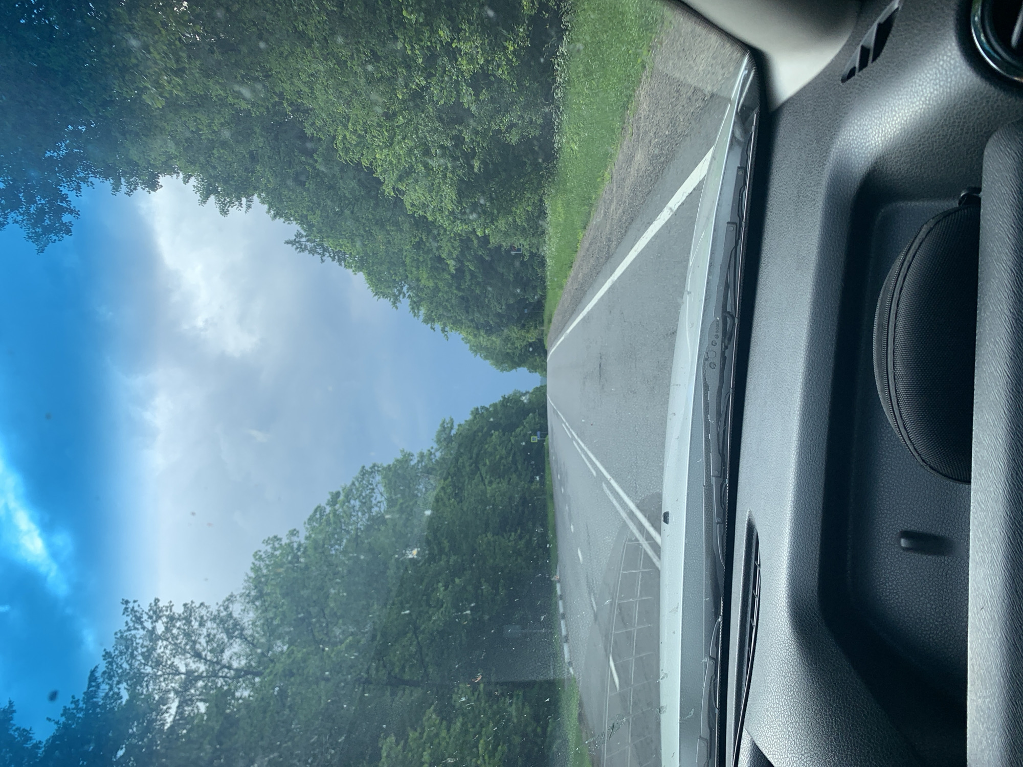The image is taken from inside a vehicle, looking forward through the windshield. The scene depicts a paved two-lane road with white markings, curving slightly to the left as it recedes into the distance. Both sides of the road are lined with dense green trees. The sky above is overcast with dramatic, bluish-gray clouds, hinting at impending rain. The dashboard of the vehicle is visible in the foreground, with a dark textured surface. In the distance, a few road signs can be observed on the right side of the road. The overall environment suggests a rural setting in Belarus, possibly near Грозаўскі сельскі Савет.