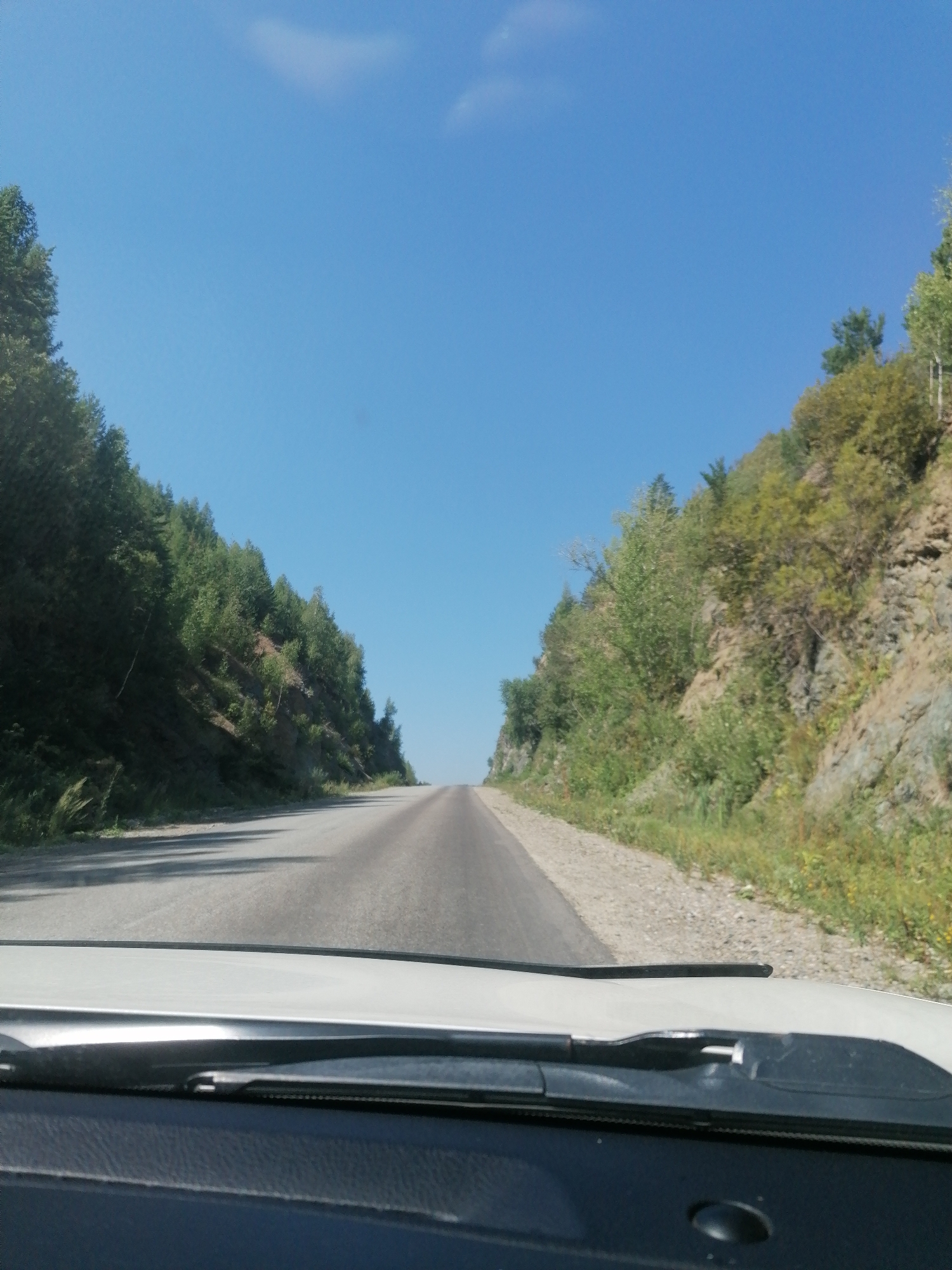 A POV shot from inside a vehicle on a sunny day, looking out at a paved road that curves slightly to the right and recedes into the distance. The road is flanked by steep, rocky hillsides covered in sparse green vegetation and trees. The sky is a clear, bright blue with a single wispy cloud. The windshield and dashboard of the vehicle are visible in the foreground. The location is identified as Tarkhanka rural district, Kazakhstan.
