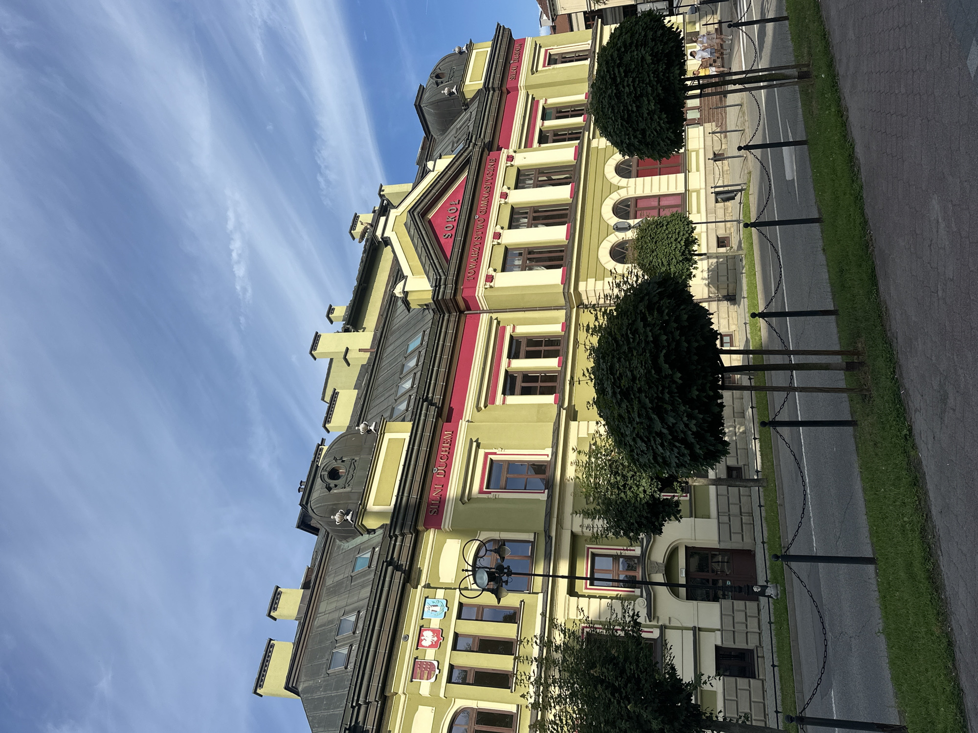 The image shows a building in Nowy Sącz, Poland, with the inscription "Sokół Towarzystwo Gimnastyczne" visible. The building is painted in light green and has red accents. The architecture features prominent chimneys, arched windows, and a domed roof section. In front of the building, there is a street with a sidewalk, green grass, and several trees, some of which are shaped into spheres. A chain fence separates the sidewalk from the street. In the background, two people are walking on the sidewalk. The sky is blue with scattered white clouds.