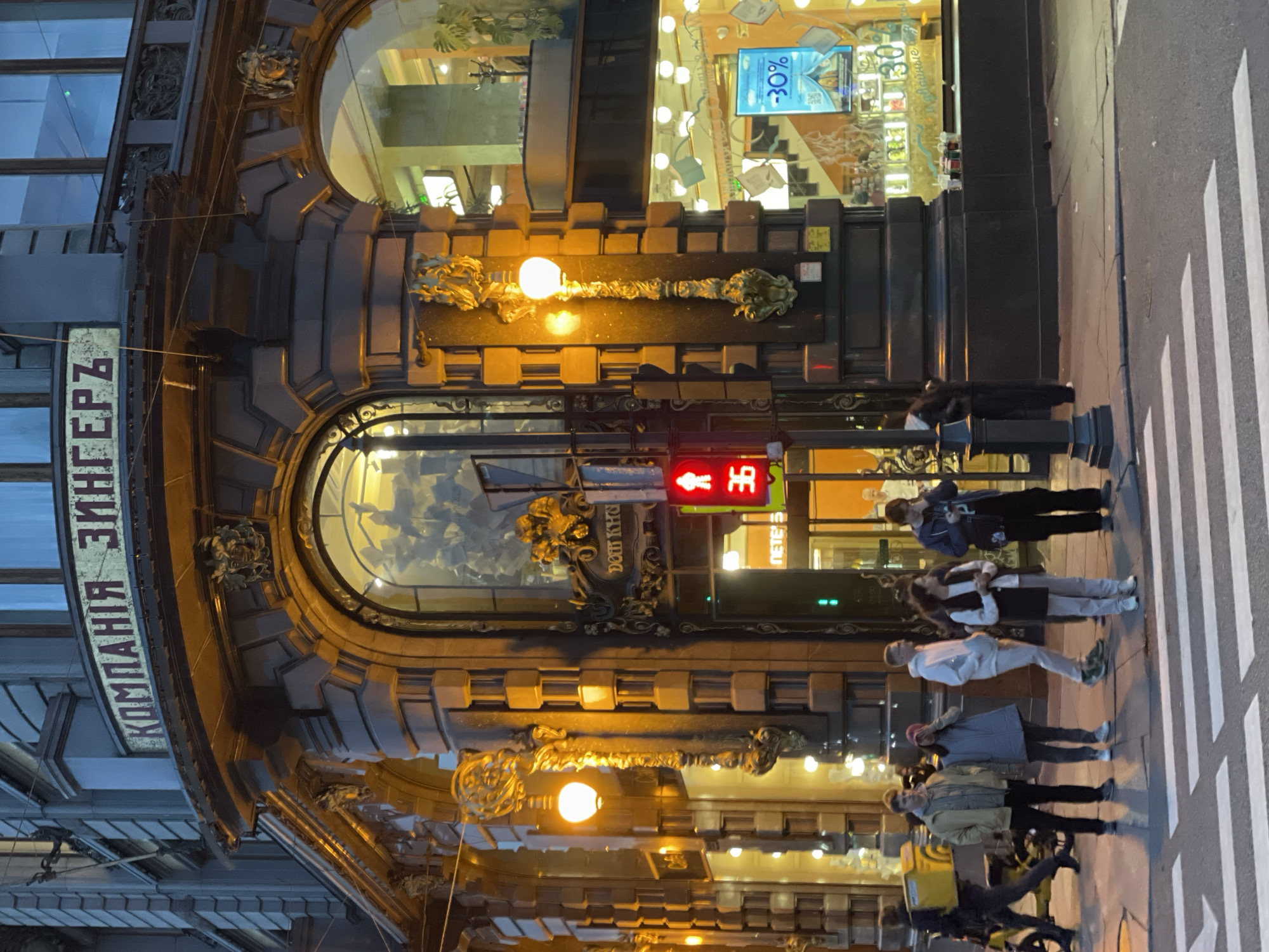 The image shows an outdoor urban scene at dusk or night, with a group of people standing on a sidewalk near a crosswalk. The building behind them features ornate architectural details and illuminated windows.
In the foreground, a crosswalk with white stripes is visible on asphalt. To the right of the crosswalk, a black lamppost stands with a red pedestrian traffic signal displaying a figure and the number "36".
A group of six people are present on the sidewalk. From left to right, the first person is a male wearing a light brown jacket and black pants, with dark hair. Next to him is a female wearing a gray vest over a darker garment and a pink hat. Following her is a male in a white hoodie and light-colored pants. To his right is a female wearing a dark vest over a white shirt and light-colored pants. Another person, a male in a dark jacket and pants, is standing further to the right, holding a phone. There is a sixth person further back, obscured.
Behind the people, the facade of a building is visible. The building has large arched windows, some of which are illuminated, revealing interiors with displays and plants. Decorative golden elements and stone carvings adorn the building's exterior. A prominent sign above one of the windows reads "КОМПАНИЯ ЗИНГЕРЪ". The lighting in the scene is a mix of warm yellow from streetlights and internal building lights, and the cooler tones of the twilight sky.
To the far left, a person on a yellow bicycle with a yellow delivery box is moving away from the camera.