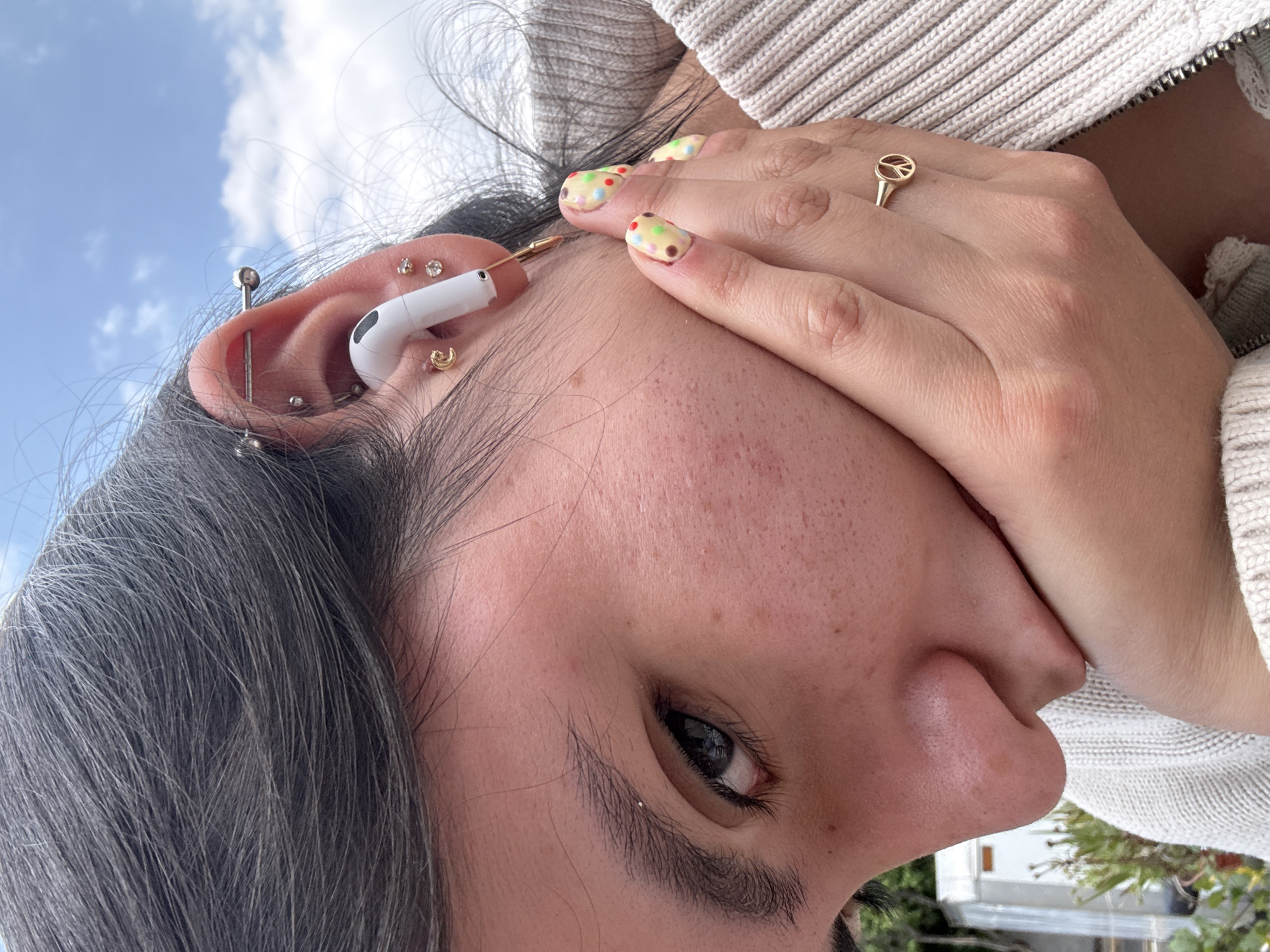 A close-up shot depicts a person with graying hair and light skin. Their left ear features multiple piercings, including a helix piercing with a barbell, two stud earrings, a tragus piercing with a crescent moon-shaped earring, and a conch piercing. An Apple AirPod Pro is visible in the ear. The person's left eye is open, and freckles are noticeable on their face. Their right hand, adorned with a gold ring featuring a peace symbol, is partially covering their mouth. The fingernails have a light yellow base with colorful polka dot designs. The person is wearing a light-colored, zipped sweater. The background shows a blurred view of a blue sky with clouds, trees, and a glimpse of a white vehicle or building. No definitive evidence indicates the location as Metepec, Mexico, but the presence of flora suggests an outdoor setting.