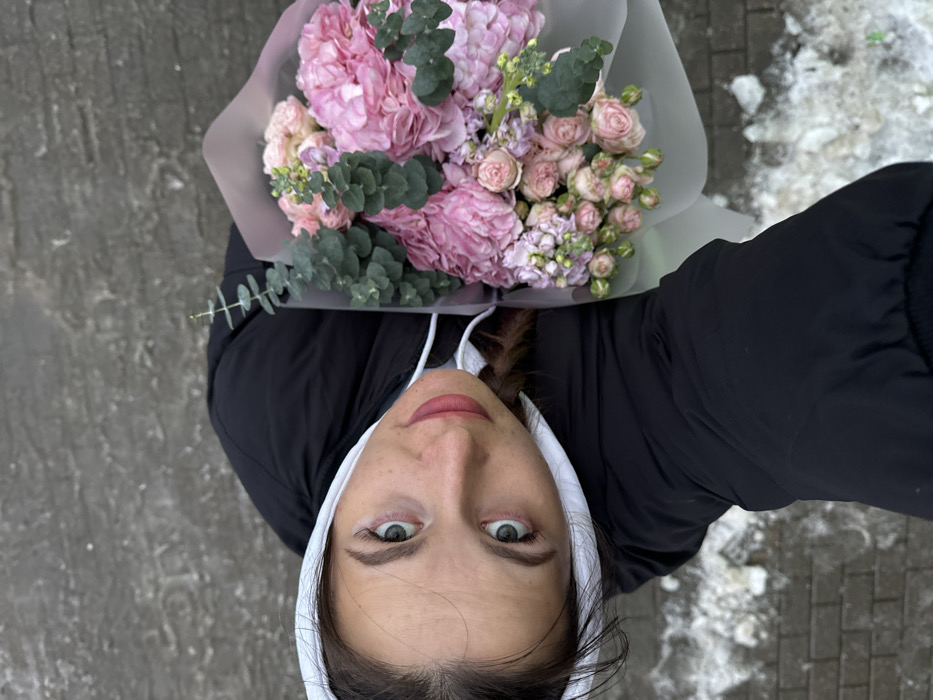 A young woman is depicted from a high-angle perspective, looking directly upwards into the camera, which she appears to be holding. She has light-colored eyes and wears a black puffer jacket over a white hooded sweatshirt. Her lips are adorned with reddish-pink lipstick. In her left arm, she holds a large bouquet of flowers wrapped in light-colored paper. The bouquet consists predominantly of pink blooms, including large hydrangeas, smaller roses or spray roses, and green eucalyptus foliage. The background shows a dark, textured ground surface, possibly asphalt or concrete, with visible patches of melting snow and slush. The specific location within the city of Odintsovo, Russia, cannot be determined from the visual information present in the image.
