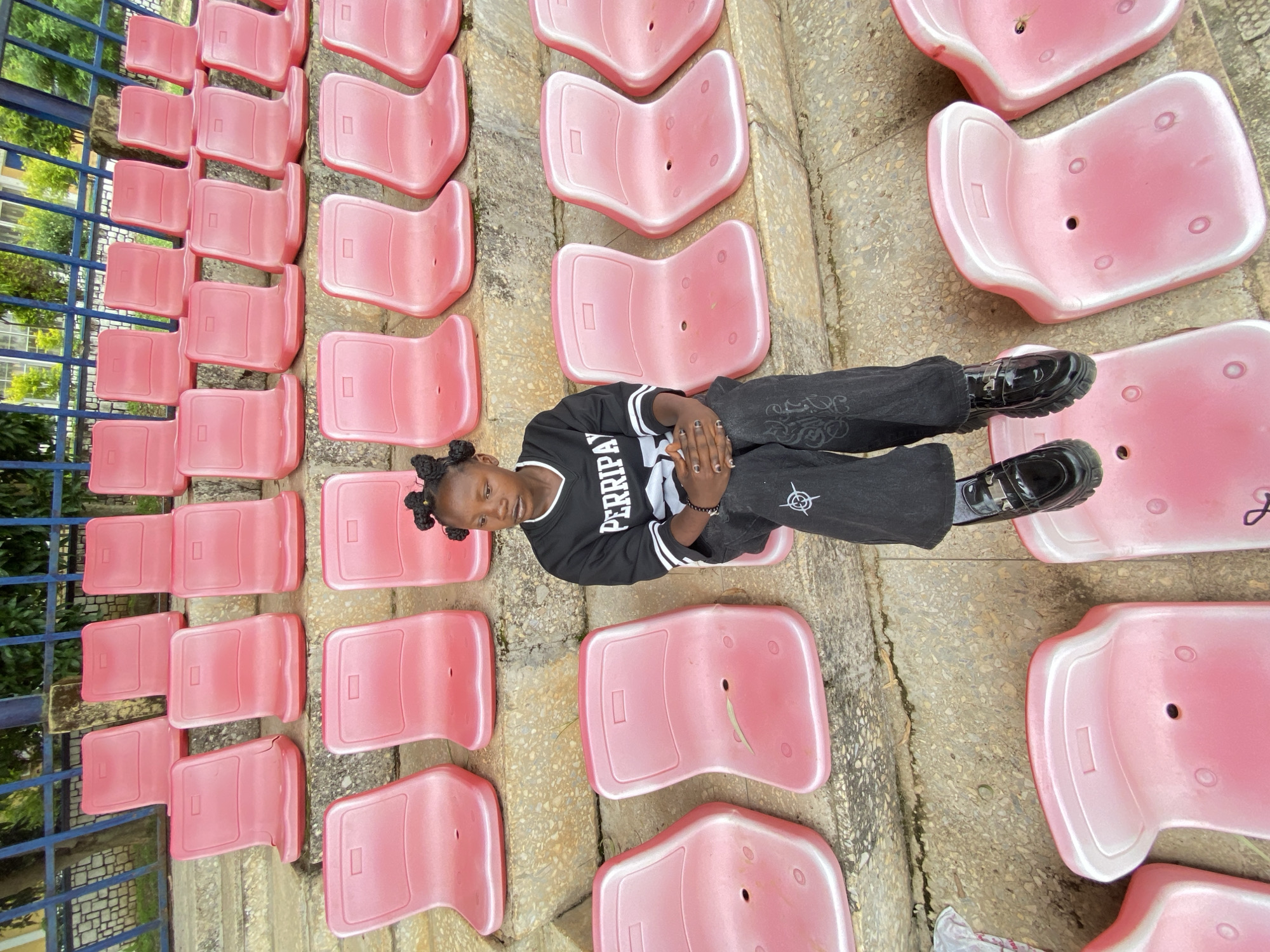 A young girl with dark skin and braided hair sits on tiered pink plastic stadium seats in Zawan, Nigeria. She is wearing a black long-sleeved shirt with white lettering that reads "PERRIPAY" and black pants with white embroidery. Her black patent leather shoes have thick soles. The stadium seats are arranged in multiple rows on a concrete or stone stepped platform. In the background, a blue metal fence separates the seating area from a path and some greenery. The overall lighting suggests it is daytime, possibly overcast.