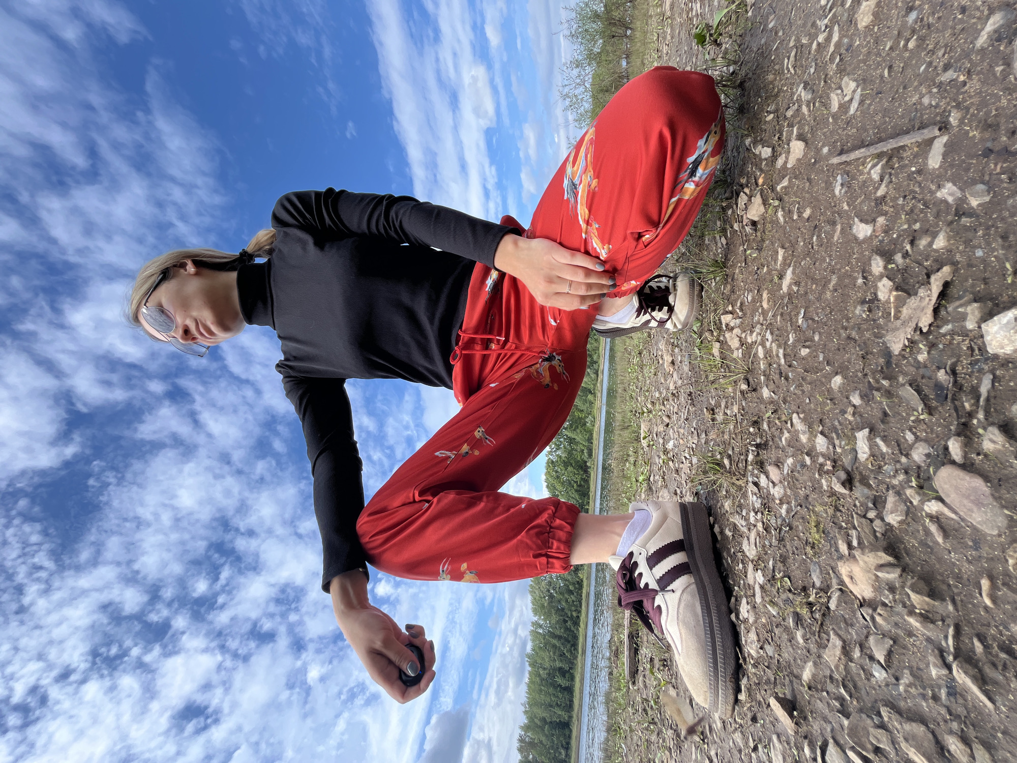 A young adult female is crouched on a pebbly shoreline, with a body of water and a tree line in the background under a cloudy blue sky. She is wearing a black turtleneck, red patterned pants, and athletic shoes. Her sunglasses are perched on her nose, and her blonde hair is pulled back in a ponytail. She is holding a small, dark object in her left hand, with her right hand resting on her knee. The scene appears to be a natural setting, likely a park or recreational area, possibly along the Kama River in Chaikovsky, Russia.