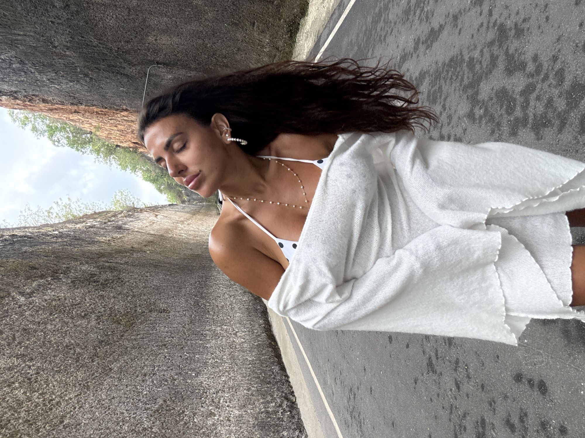 A woman stands on a paved road between two sheer rock walls in Kuta Selatan, Indonesia. She is wearing a white polka dot bikini top and a white loosely draped cardigan. Her eyes are closed, and her head is tilted back as if basking in sunlight. She has long, dark hair and is wearing pearl earrings and a delicate pearl necklace. The road has a white line painted down the center. The rock walls are rough and textured, with some vegetation growing at the top on the right side. The sky is visible between the walls.