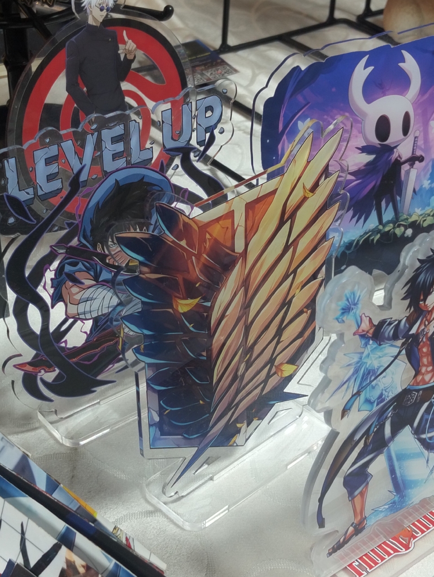 A group of anime-style acrylic figures stand on a white surface. The figure on the left depicts a man with white hair and sunglasses, wearing a dark jacket, with the words "LEVEL UP" prominently displayed. Next to him is a figure of a character from Hollow Knight, a white-headed creature with a dark cape and a sword. To the right of that, a character with black hair and a blue hood is partially obscured by a large, golden, wing-like object. Further to the right, a character with dark hair and an orange-toned shirt is shown with ice-like projectiles around him. The background is blurred, showing what appears to be a black metal rack.