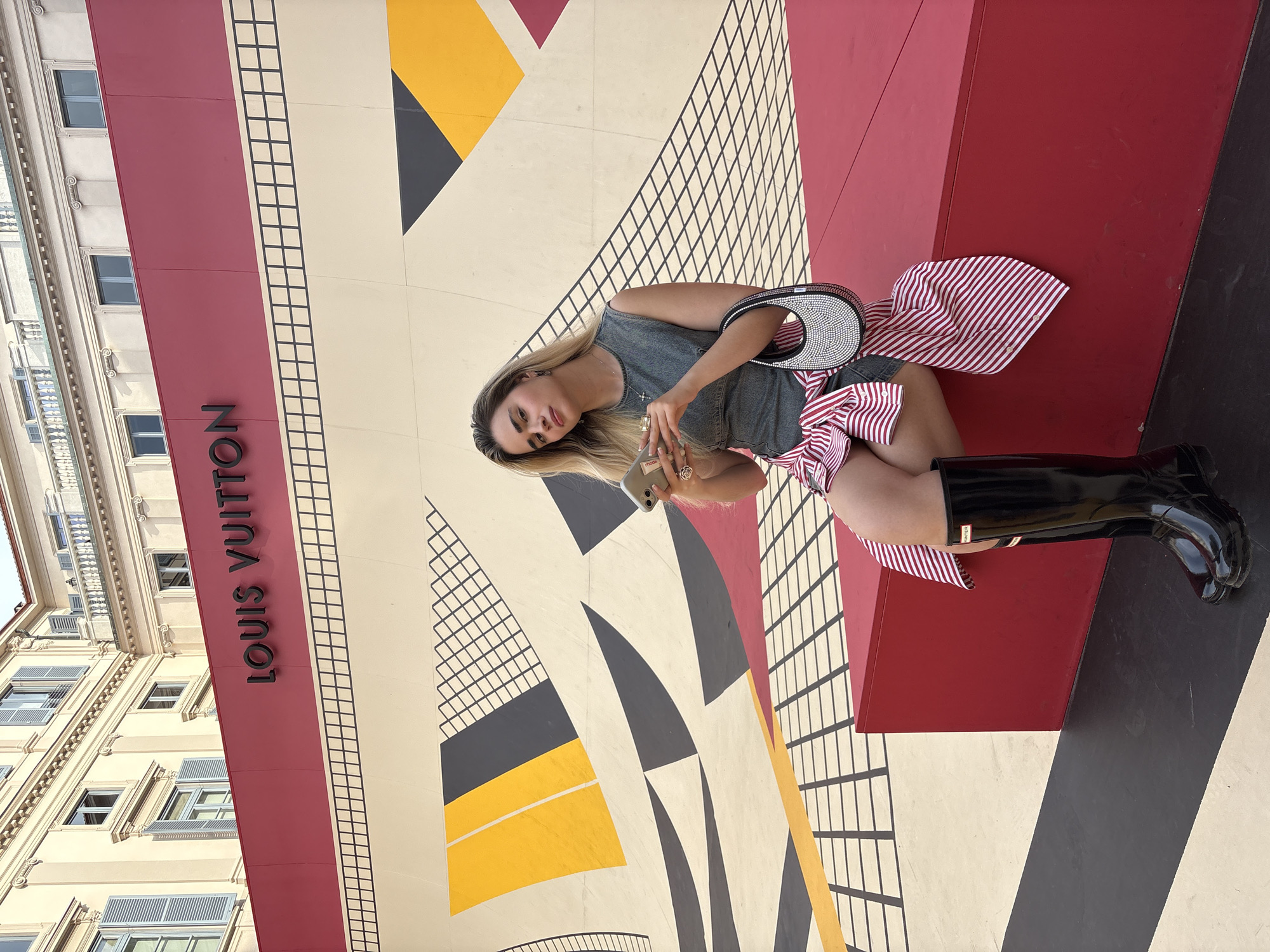 A woman is sitting on a red, black, and white geometric floor in front of a building in Milan, Italy, that features the Louis Vuitton logo. The building is a light-colored stone with multiple windows. The woman is wearing a grey dress, a red and white striped shirt, and black boots. She is holding a phone in her hand. She also has a rhinestone bag next to her. The floor has a grid pattern and geometric shapes in yellow, black, white, and red.